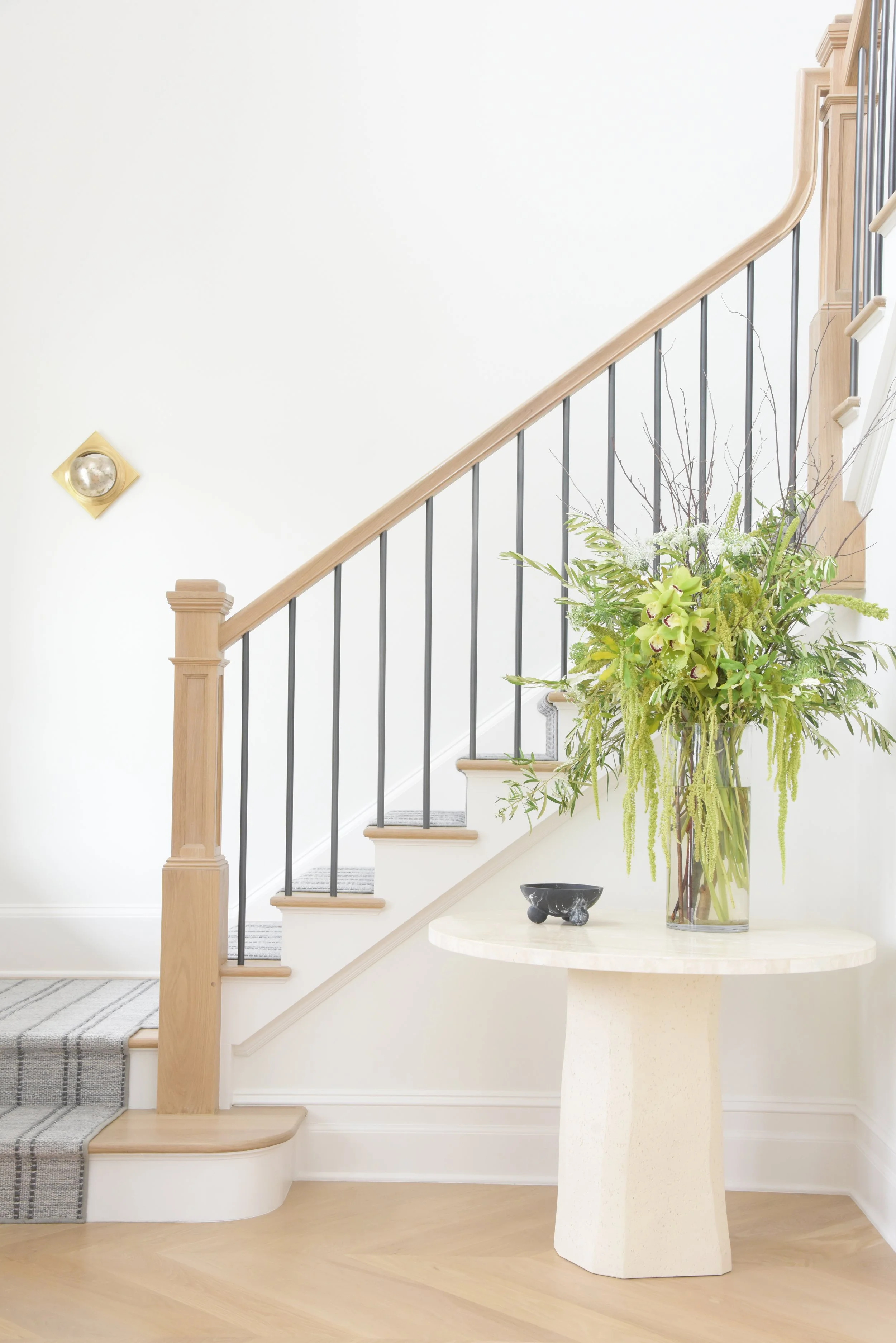 Interior view of a staircase with a wooden handrail, black metal balusters, a small gray striped rug on the steps, a round cream-colored pedestal table with a large glass vase filled with green and white flowers and greenery, and a small black decora