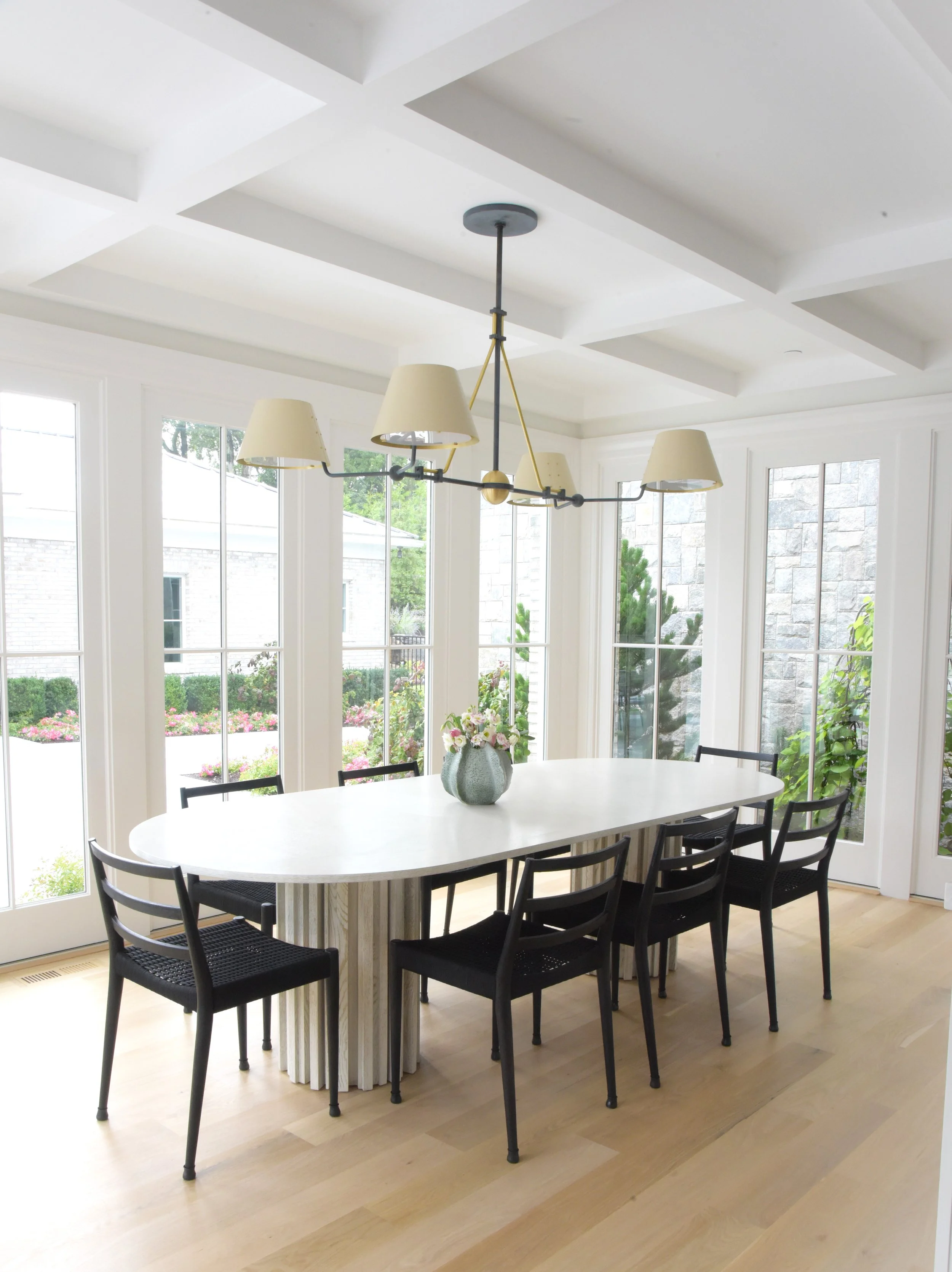 Bright dining room with a white oval table, six black chairs, a floral centerpiece, large windows, a modern chandelier, and a view of an outdoor garden.