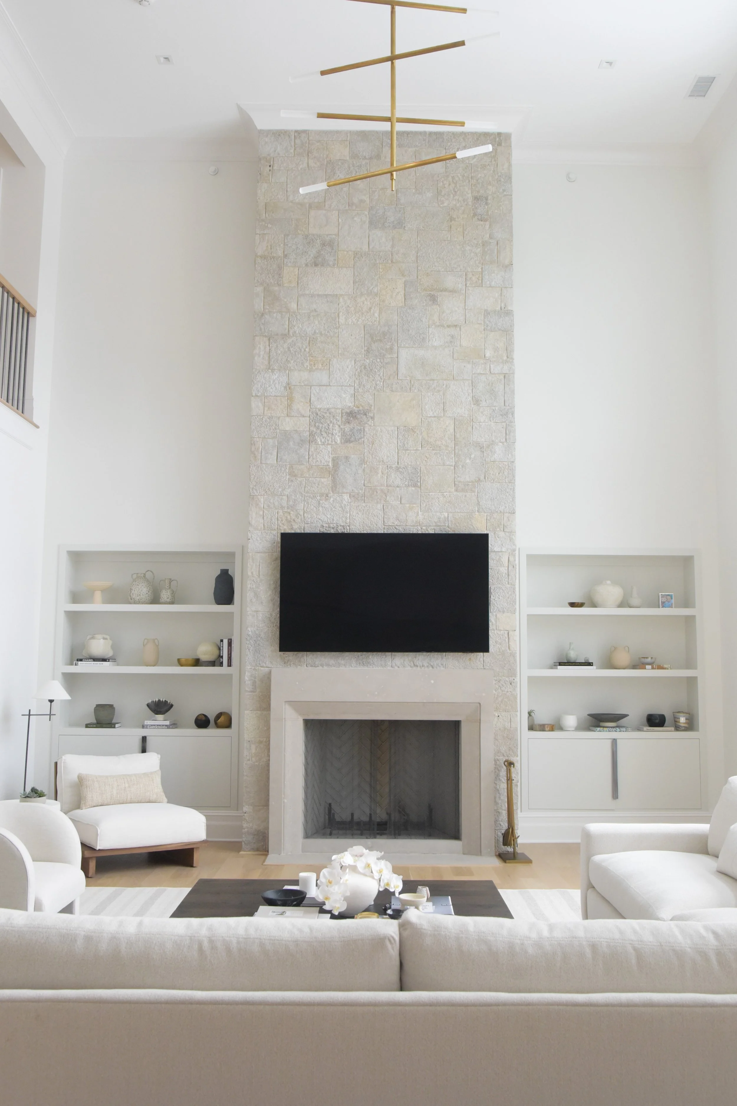 Living room with a stone fireplace, a mounted flat-screen TV, white sofas, built-in shelves with decorative items, and a contemporary gold chandelier.