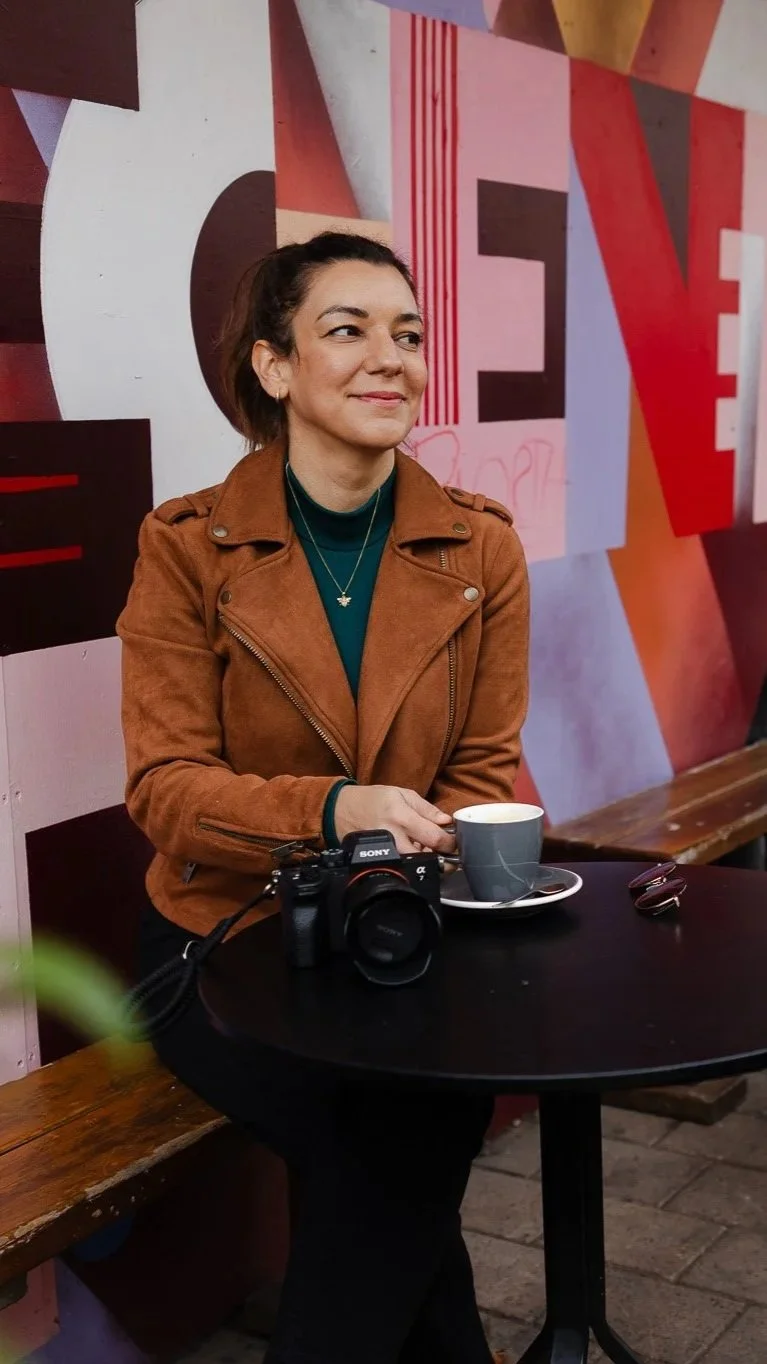 A woman sitting at a table in a coffee shop, holding a cup, with a camera placed on the table. She is wearing a brown jacket and a teal top, with a colorful geometric mural in the background.