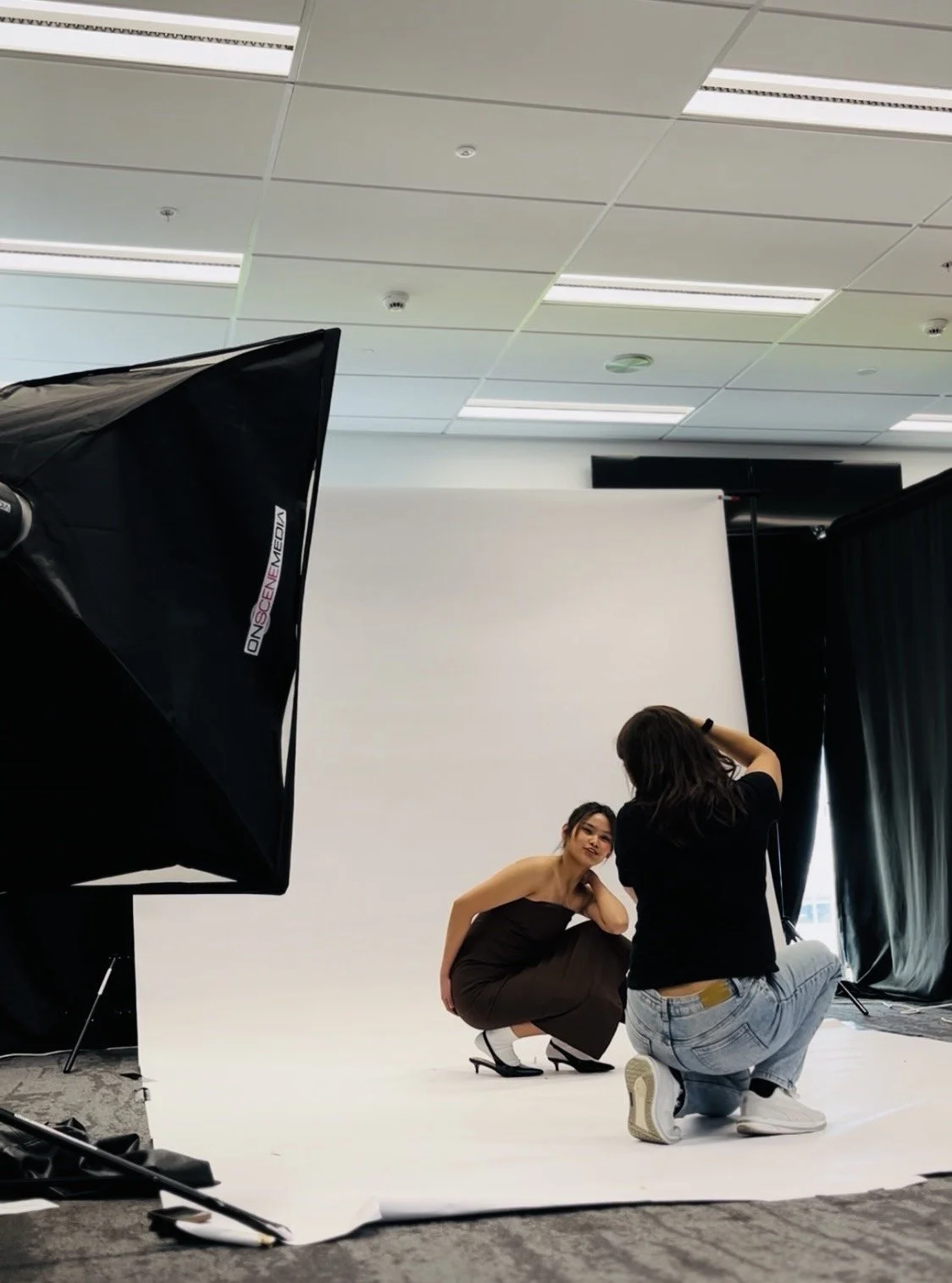 A woman in a black strapless dress and heels is squatting on a white photography backdrop, posing for a photo taken by a person kneeling and holding a camera. A large studio softbox light is visible on the left, and black curtains and a white screen are in the background.