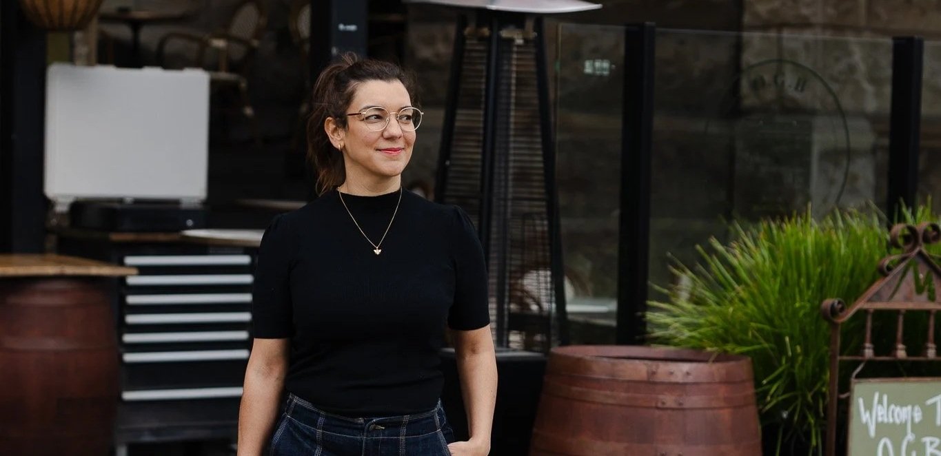 A woman with short dark hair, glasses, and a black top standing outside near a plant and a chalkboard sign that says "Welcome".