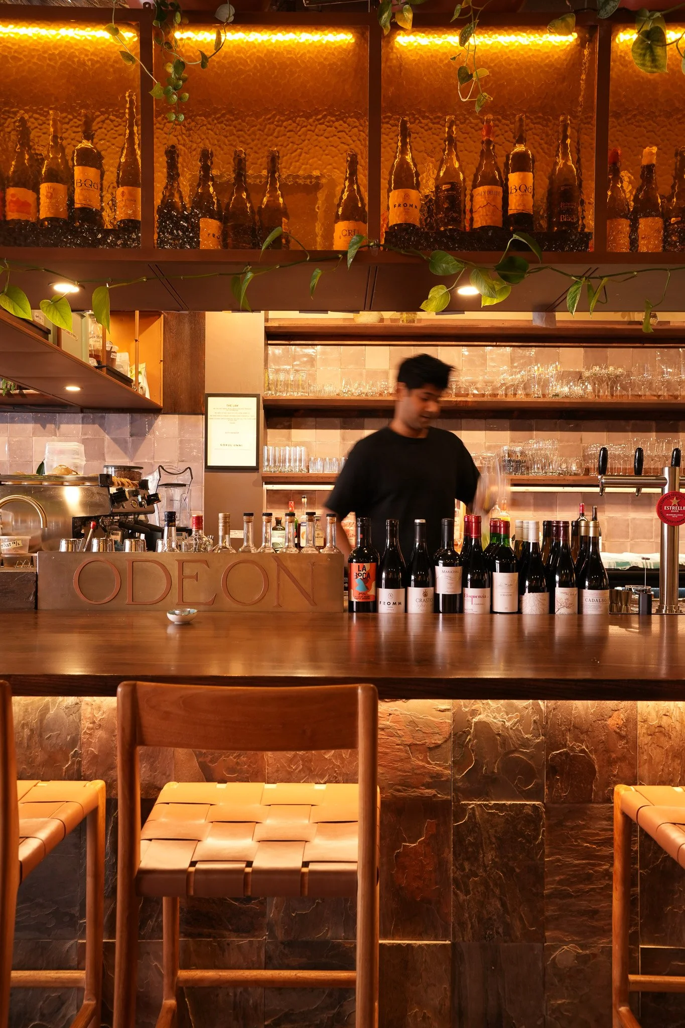 A bar scene with a man standing behind the counter, bottles of wine lined up in front, glassware on shelves behind, warm lighting, wood and stone decor, with some chairs in the foreground.