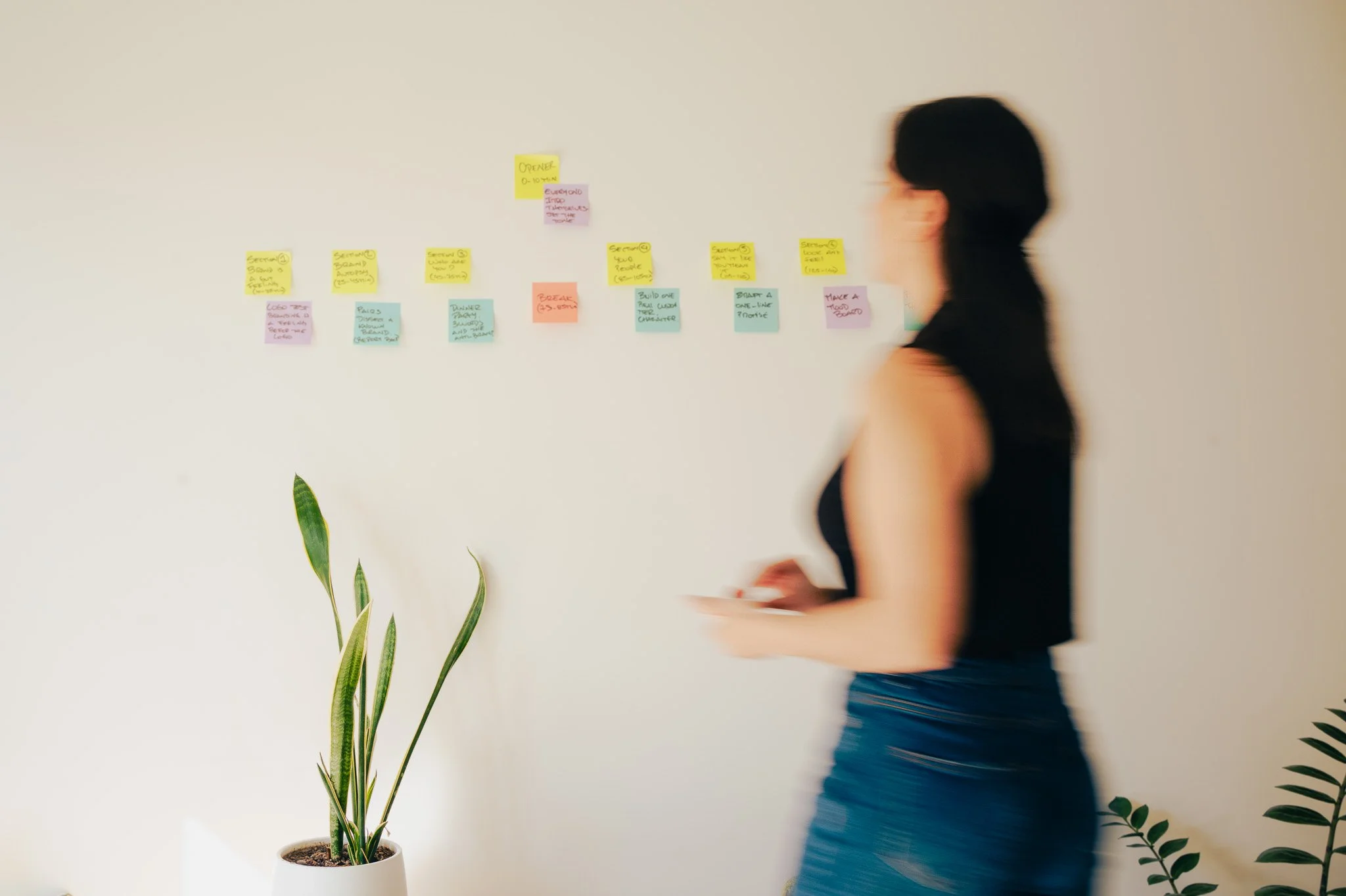 A woman walking past a white wall with colorful sticky notes arranged in a line. A potted plant with long green leaves is in the foreground.