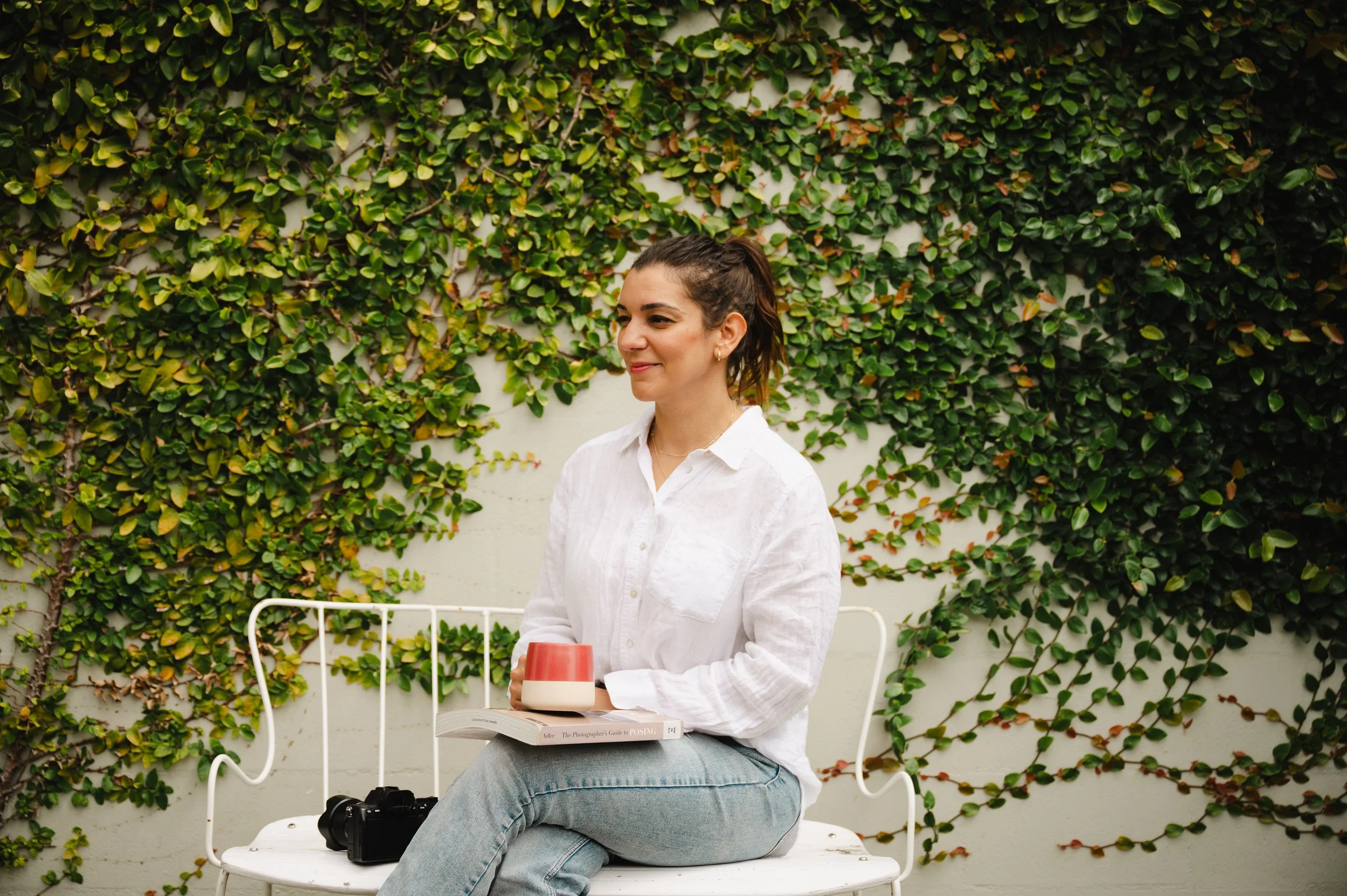 A woman with brown hair tied back, wearing a white shirt and light blue jeans, sitting on a white bench in front of a green vine-covered wall. She has a red and white ceramic mug in her left hand, an open book and a camera on her lap, and appears to be smiling slightly.