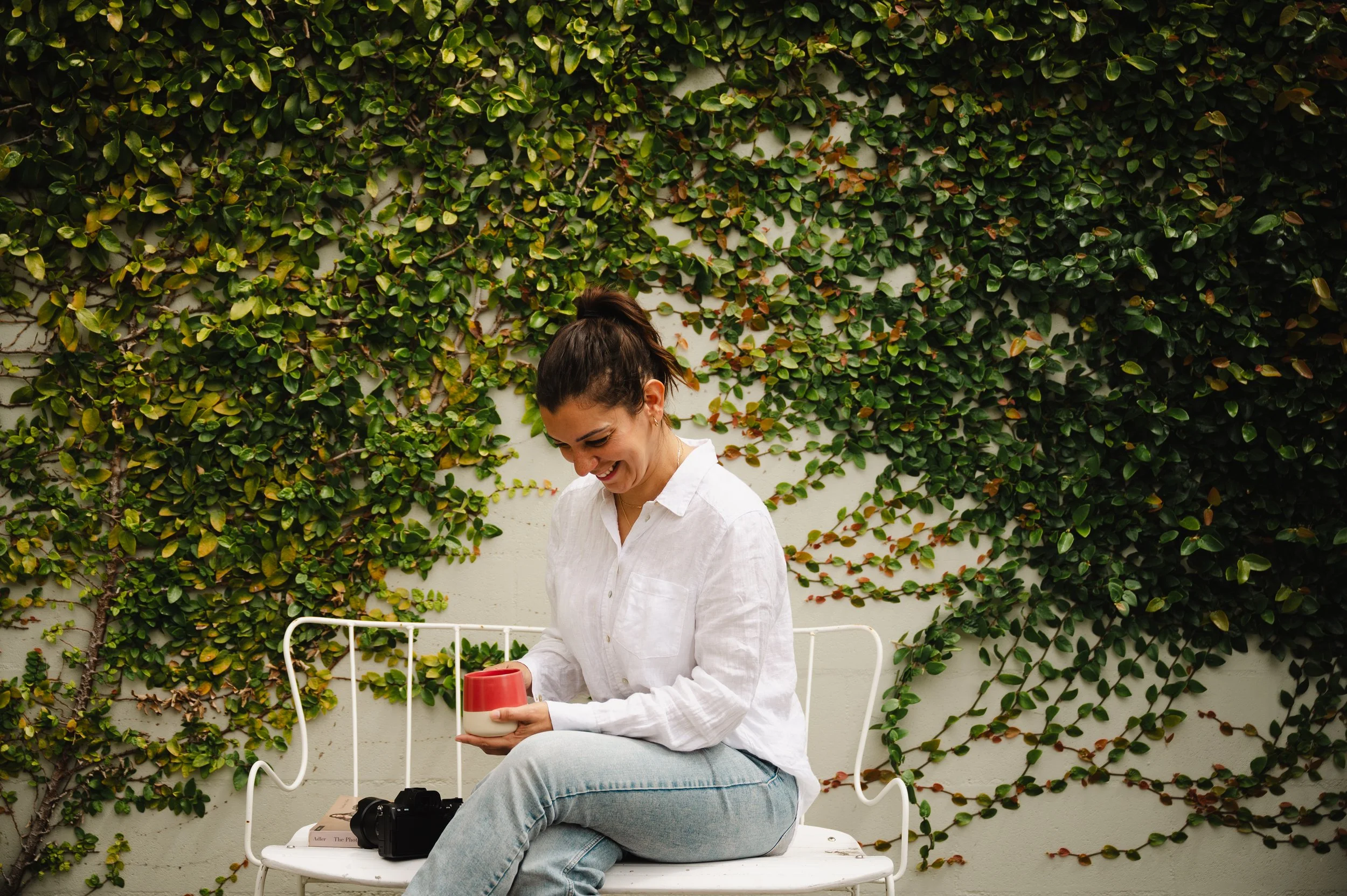 A woman with dark hair in a ponytail, wearing a white shirt and light blue jeans, is sitting on a white metal bench in front of a wall covered with green ivy. She is smiling and holding a red and white ceramic cup. There are a camera, a couple of books, and a pair of glasses on the bench nearby.