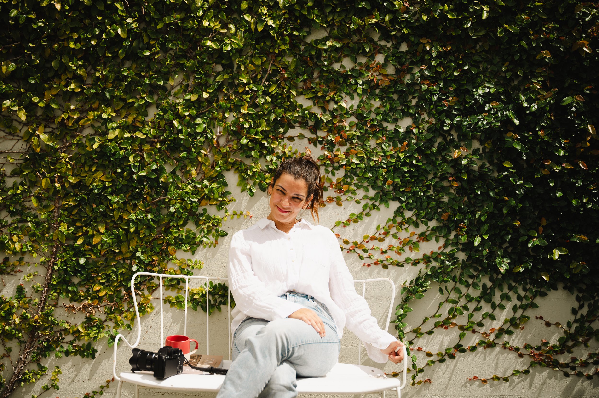 A woman sitting outdoors in front of a wall covered with green ivy, wearing a white shirt and jeans, smiling and holding a green ball, with a camera and red mug on a table nearby.