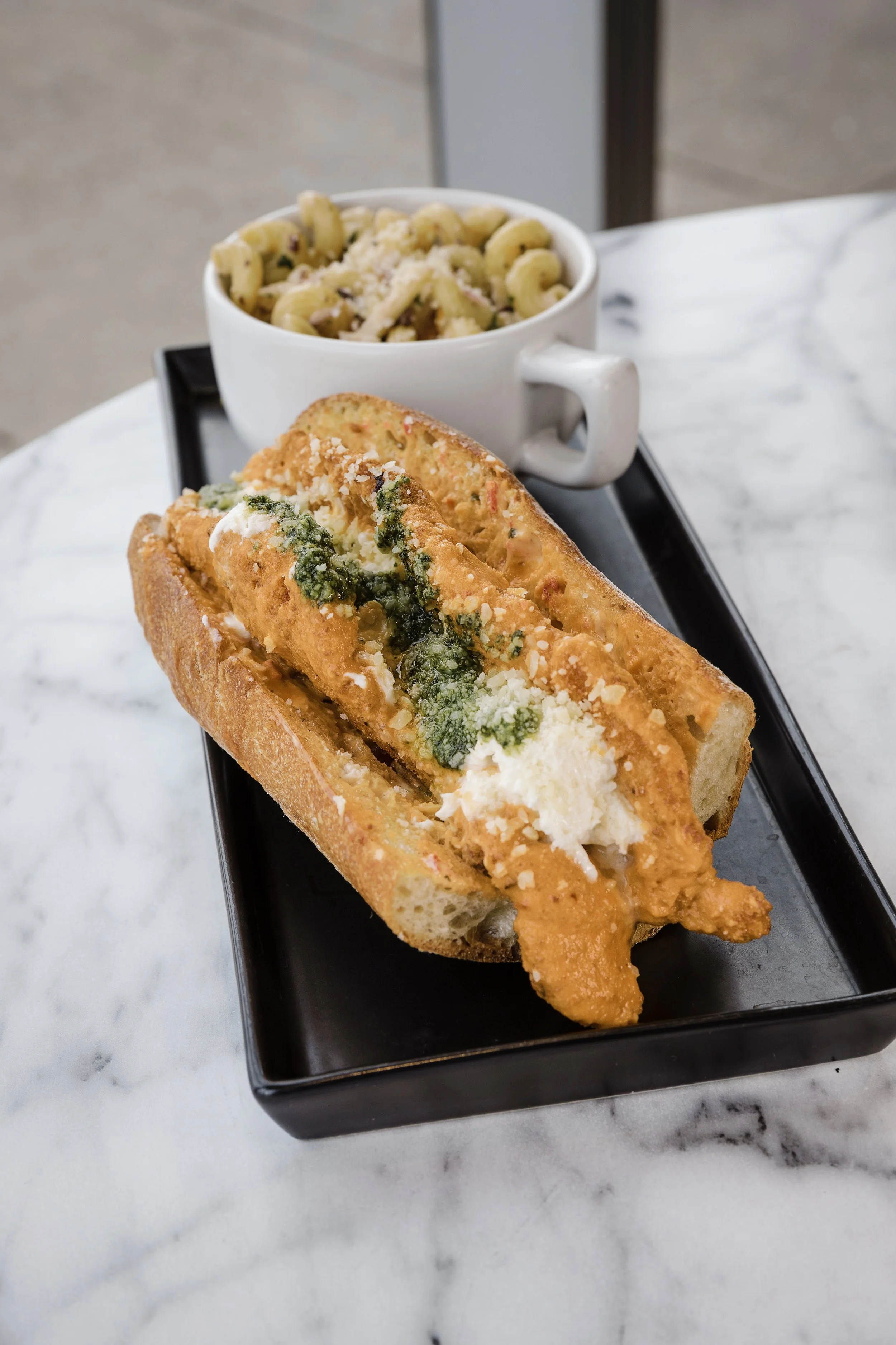 Fried fish sandwich with green sauce and cheese, served on a toasted bun, accompanied by a side of macaroni salad in a white mug on a black tray.