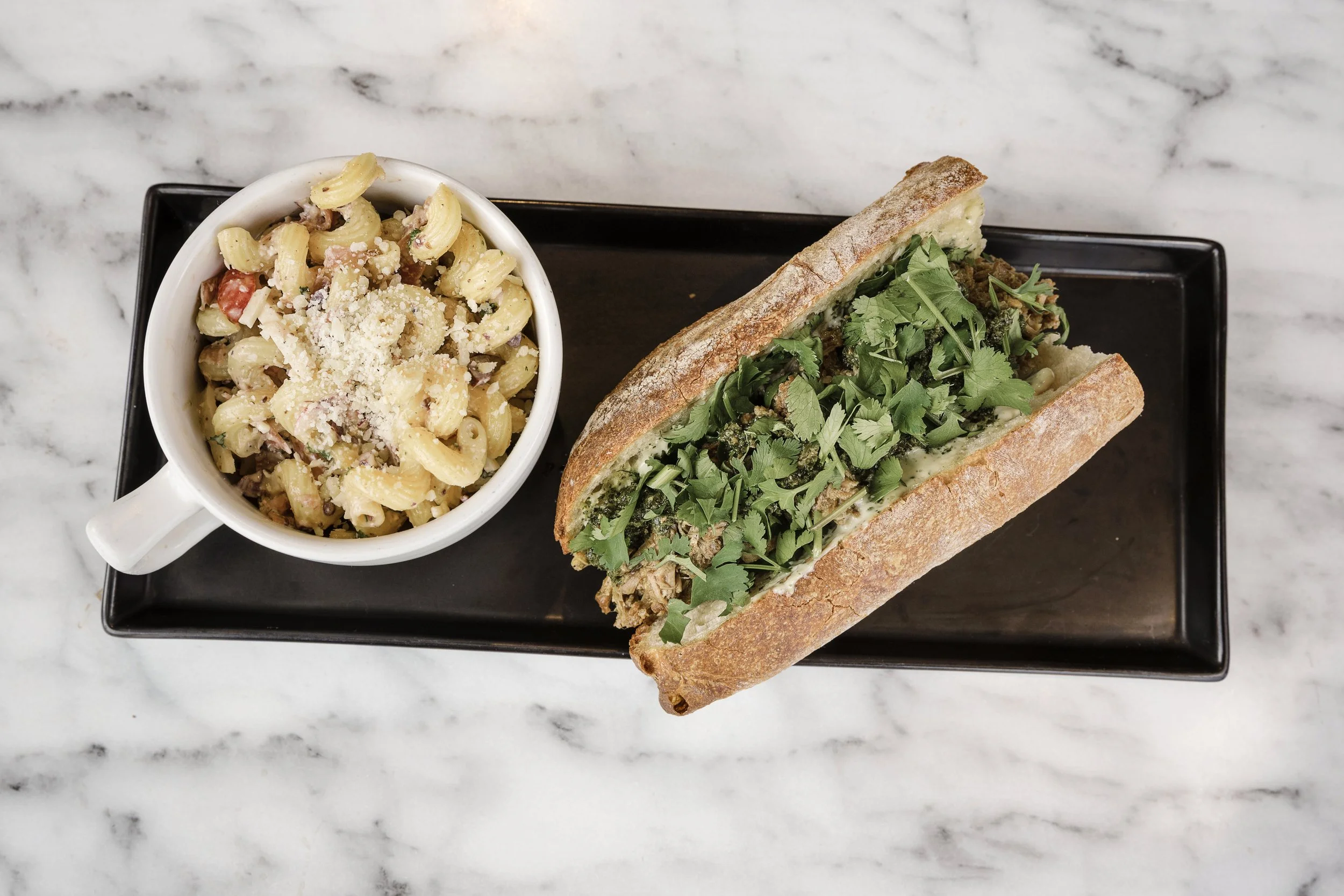 A plate with macaroni salad and a sandwich with cilantro on a black tray, marble countertop background.