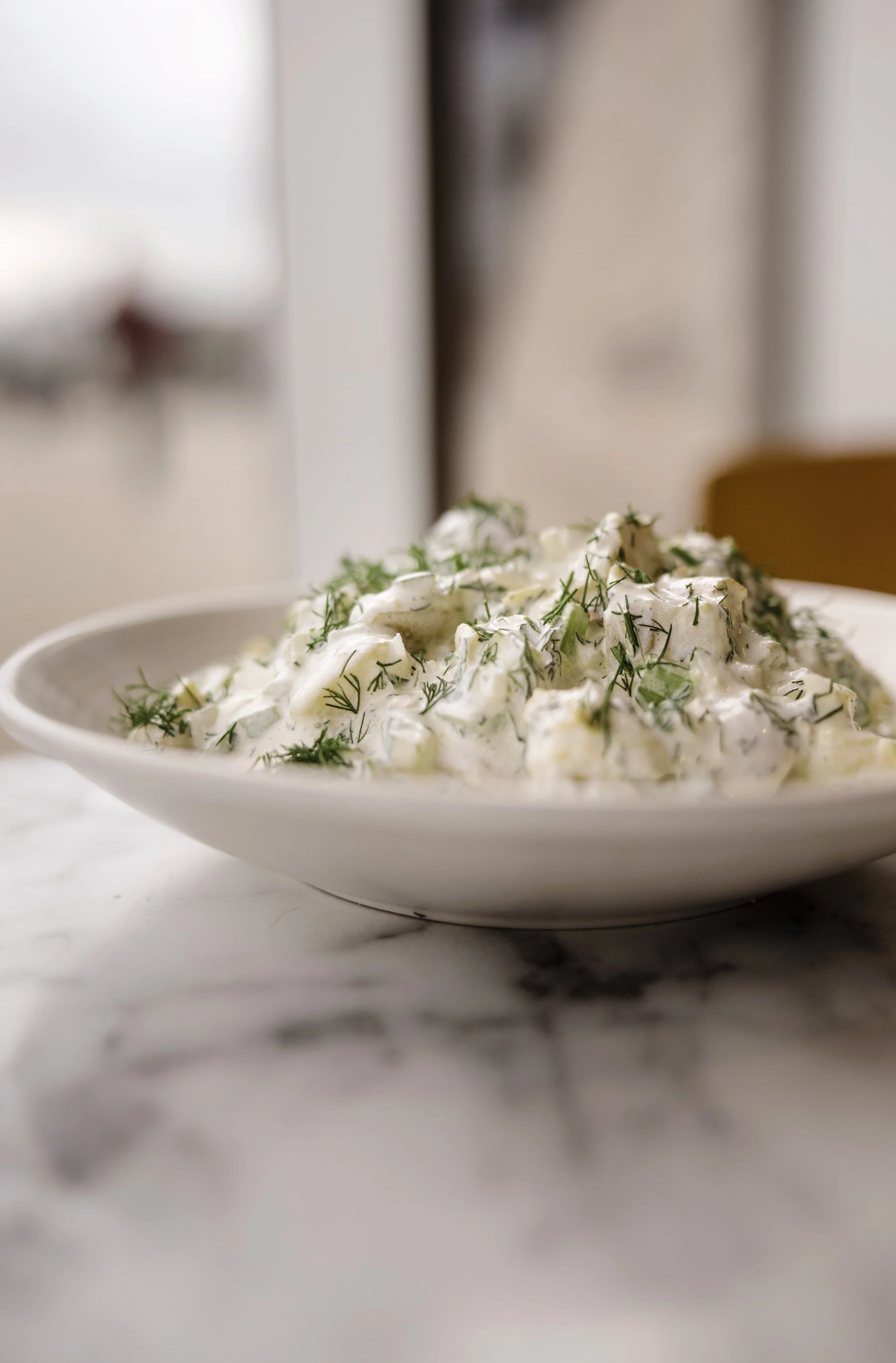 A bowl of potato salad garnished with chopped dill herbs, sitting on a marble countertop.