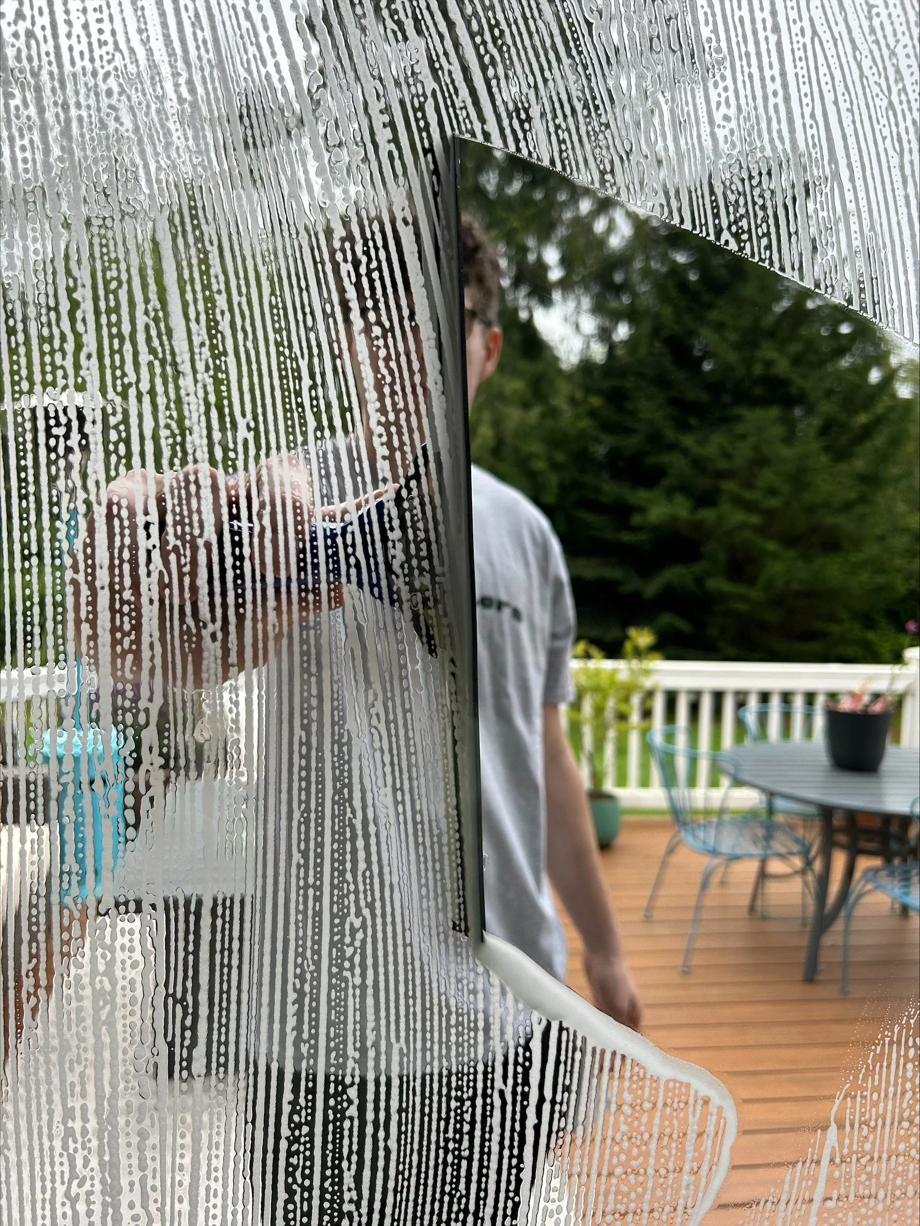 A person is seen from behind, holding a spray bottle, cleaning a glass door with water and soap. The glass has soap streaks and water droplets, and a green outdoor area with trees and a wooden deck are visible through the glass.