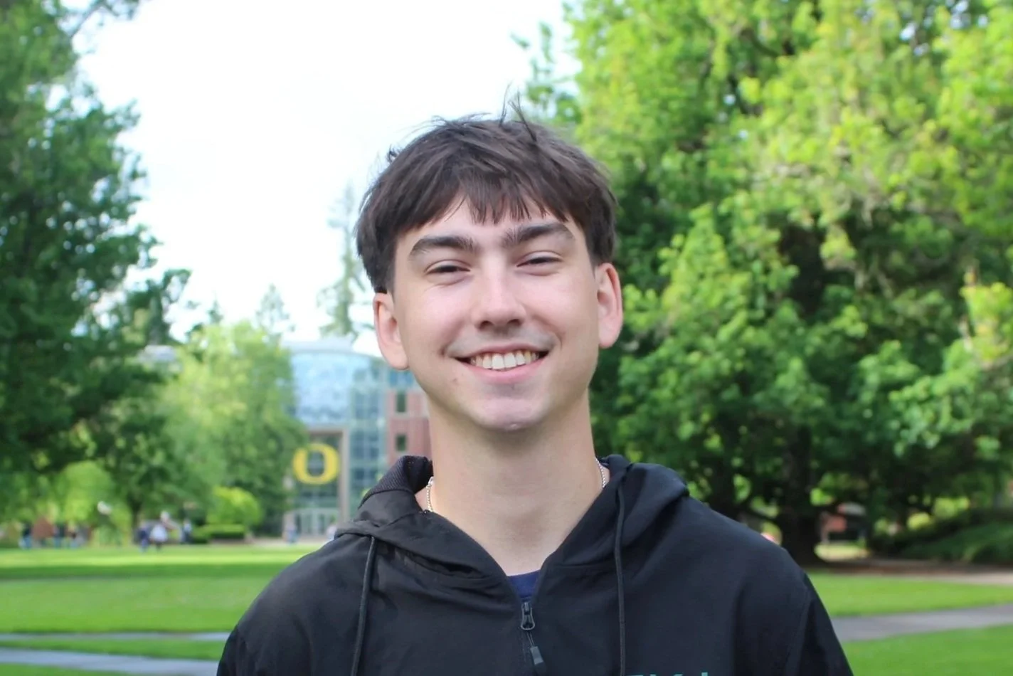 Smiling young man with brown hair wearing a black jacket outdoors in a park with green trees and a modern building in the background.