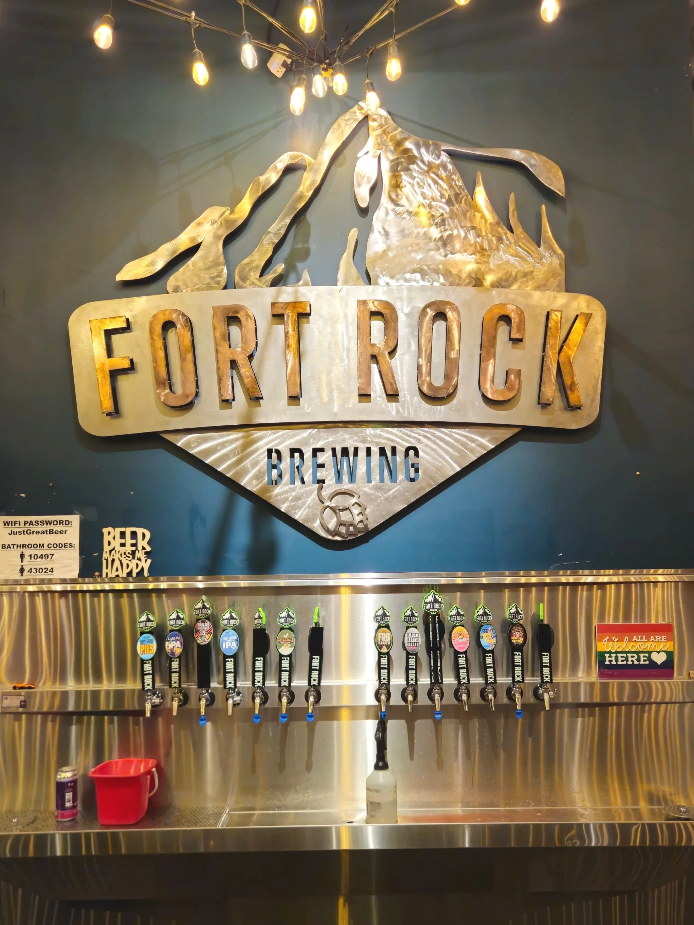 Metal sign for Fort Rock Brewing with a mountain graphic above. Multiple beer taps labeled with different beer types. Small signage indicating WiFi password, bathroom codes, and a welcome sign.