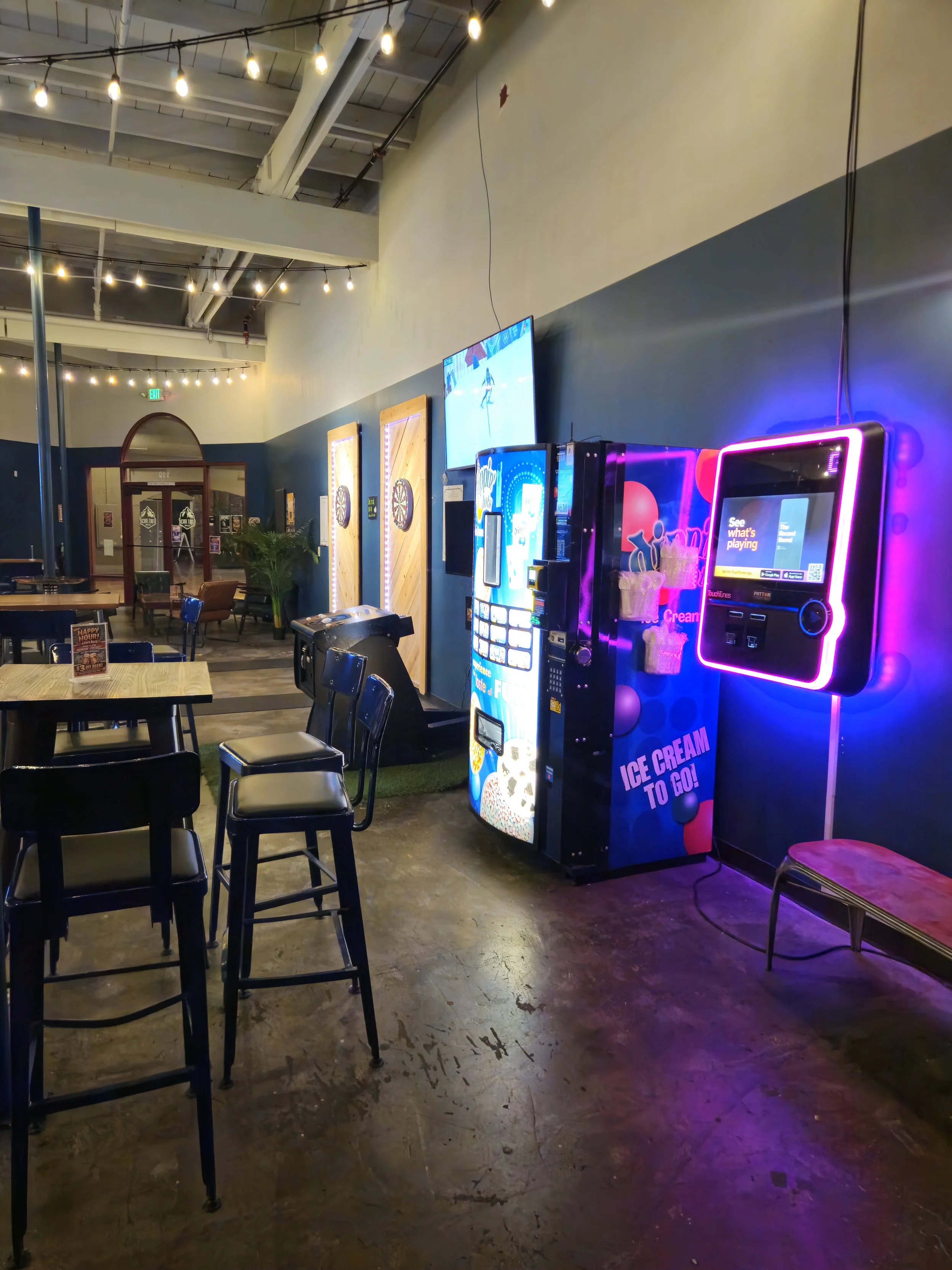 Inside a modern entertainment venue with bar seating, dart machines, vending machines, and a neon-lit touch screen kiosk.