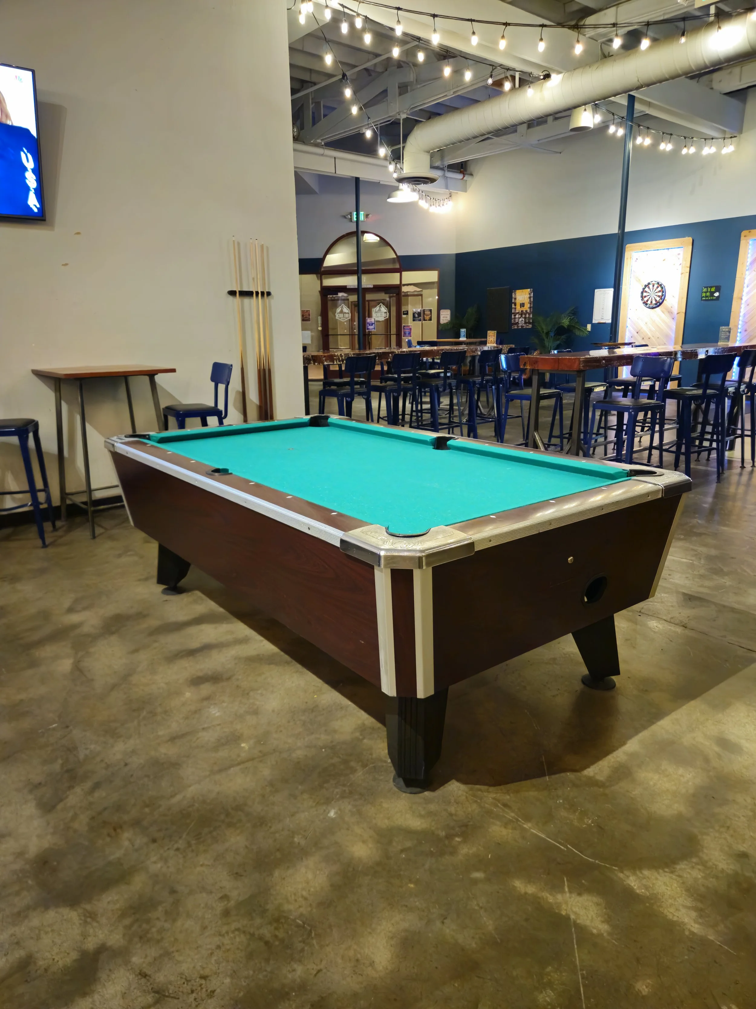 An arcade with a pool table in the foreground, bar-height tables and chairs in the background, and dartboards on the wall. String lights hang from the ceiling.
