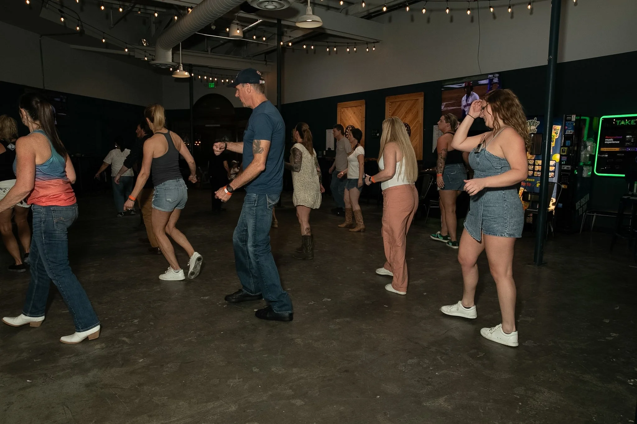 People line dance in a dimly lit indoor venue with string lights, arcade games, and a dark floor.