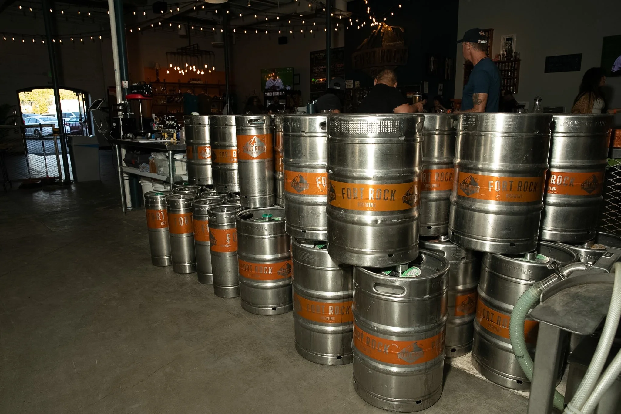 A stack of stainless steel kegs labeled 'Fort Rock Brewing' in a brewery or bar setting. There are several kegs arranged on the floor and some on a counter in the background. People are visible in the background, and the area is decorated with string