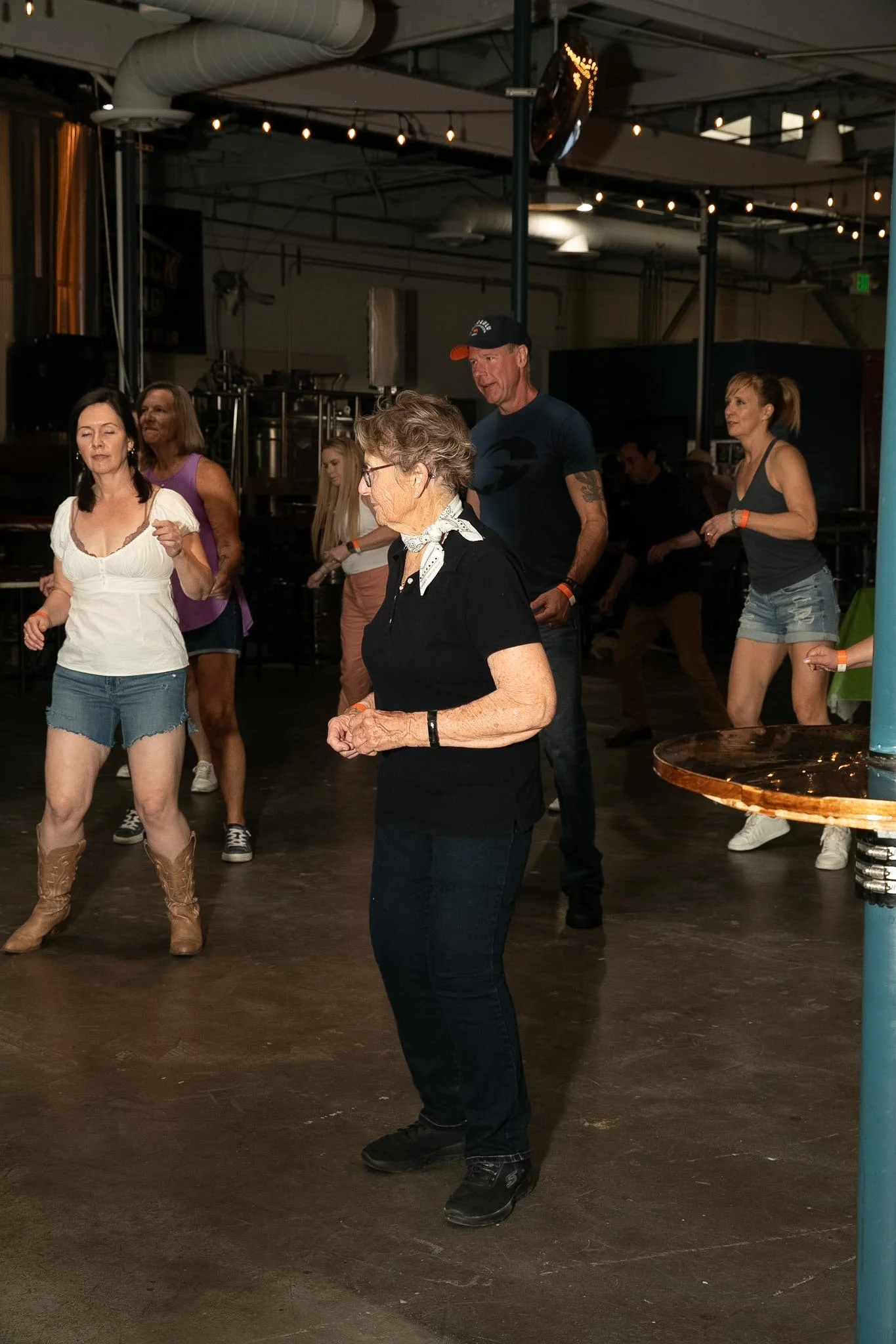 People are line dancing indoors; mostly women, one man, wearing casual country-western style clothing, in a dimly lit venue with lights hanging from the ceiling.