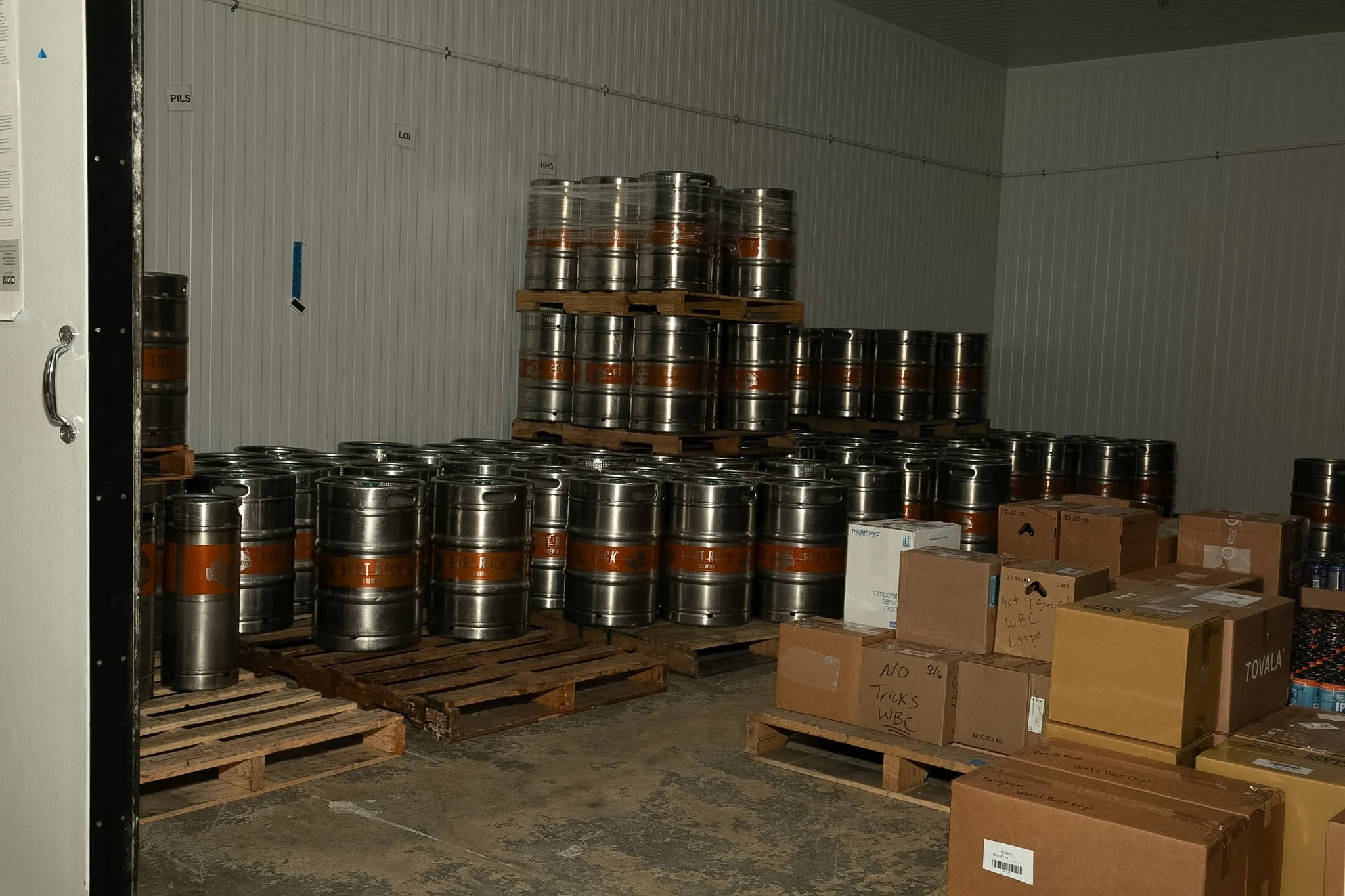 A storage room with stacks of metal kegs on pallets and cardboard boxes, some labeled with notes, against a white paneled wall.