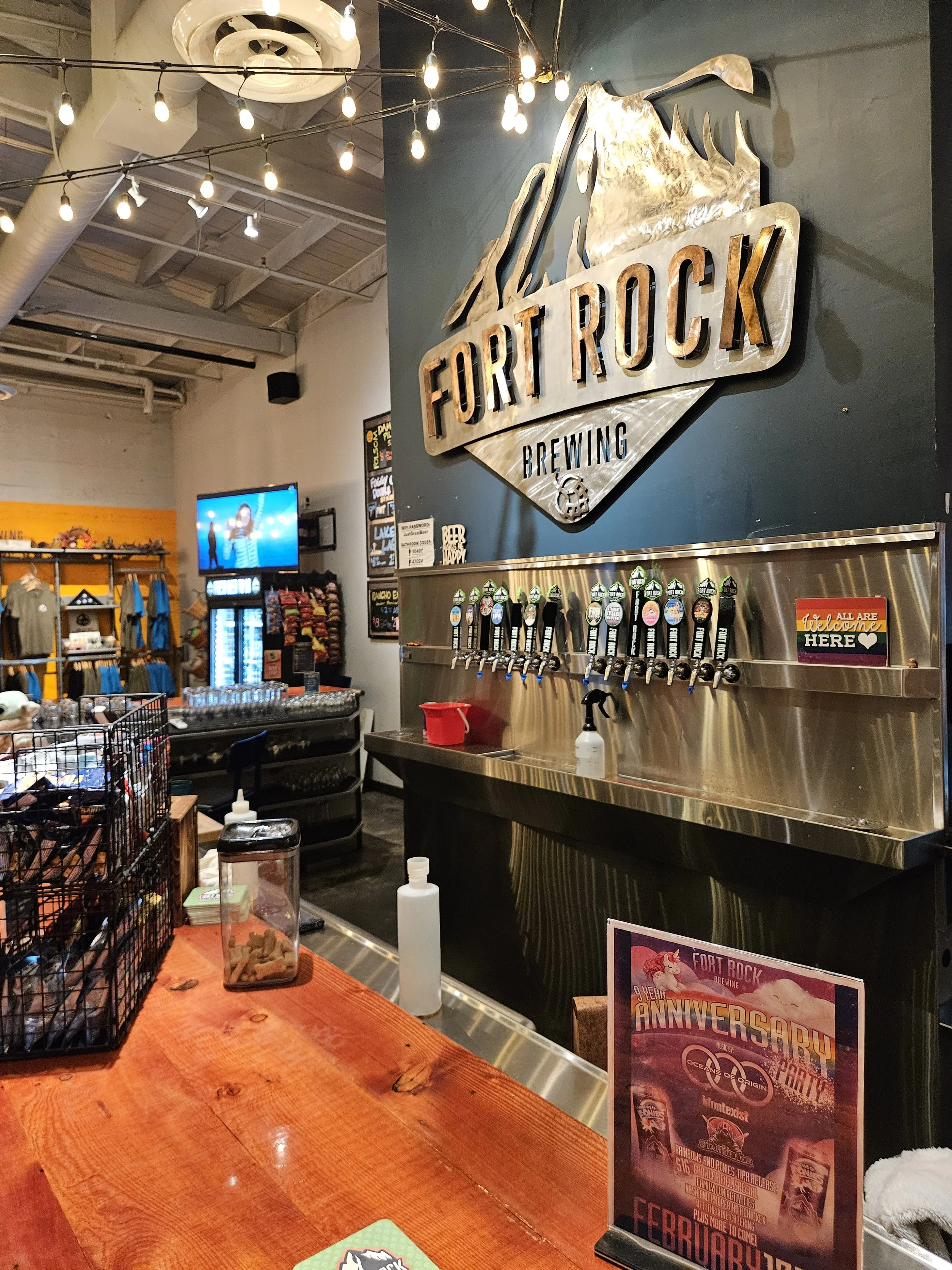Interior of Fort Rock Brewing with beer tap handles on a stainless steel wall, a large Fort Rock logo on the wall, and a bar counter with hand sanitizer, snacks, and promotional flyers. String lights hang from the ceiling.