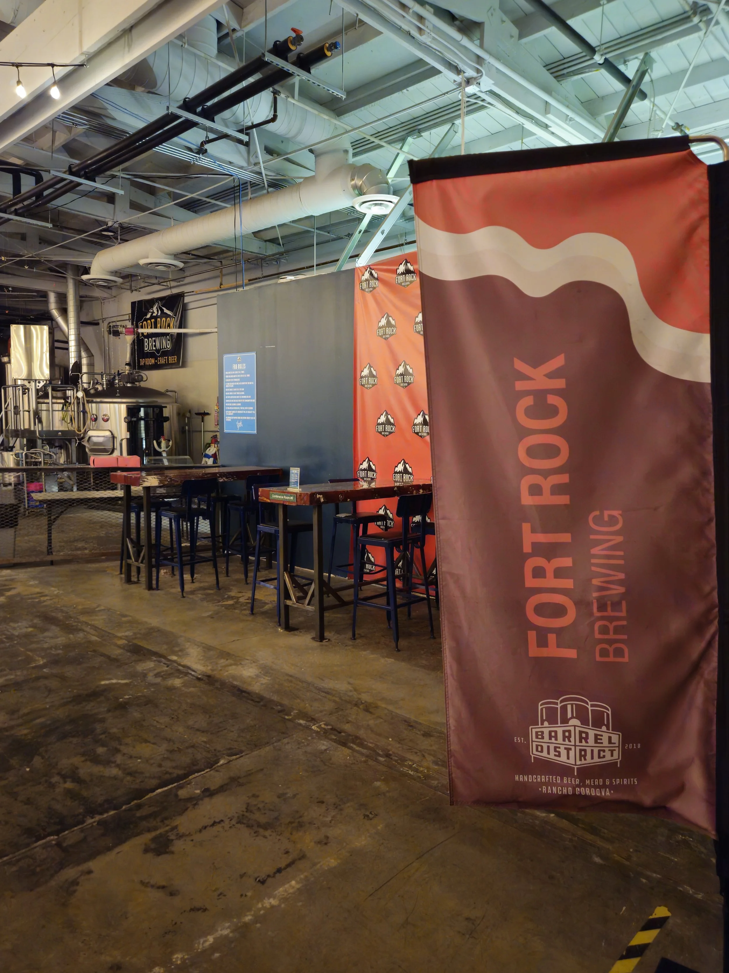 Interior of a brewery with a banner reading 'Fort Rock Brewing' and a sign 'Barrel District' on it, with brewing equipment and tables.