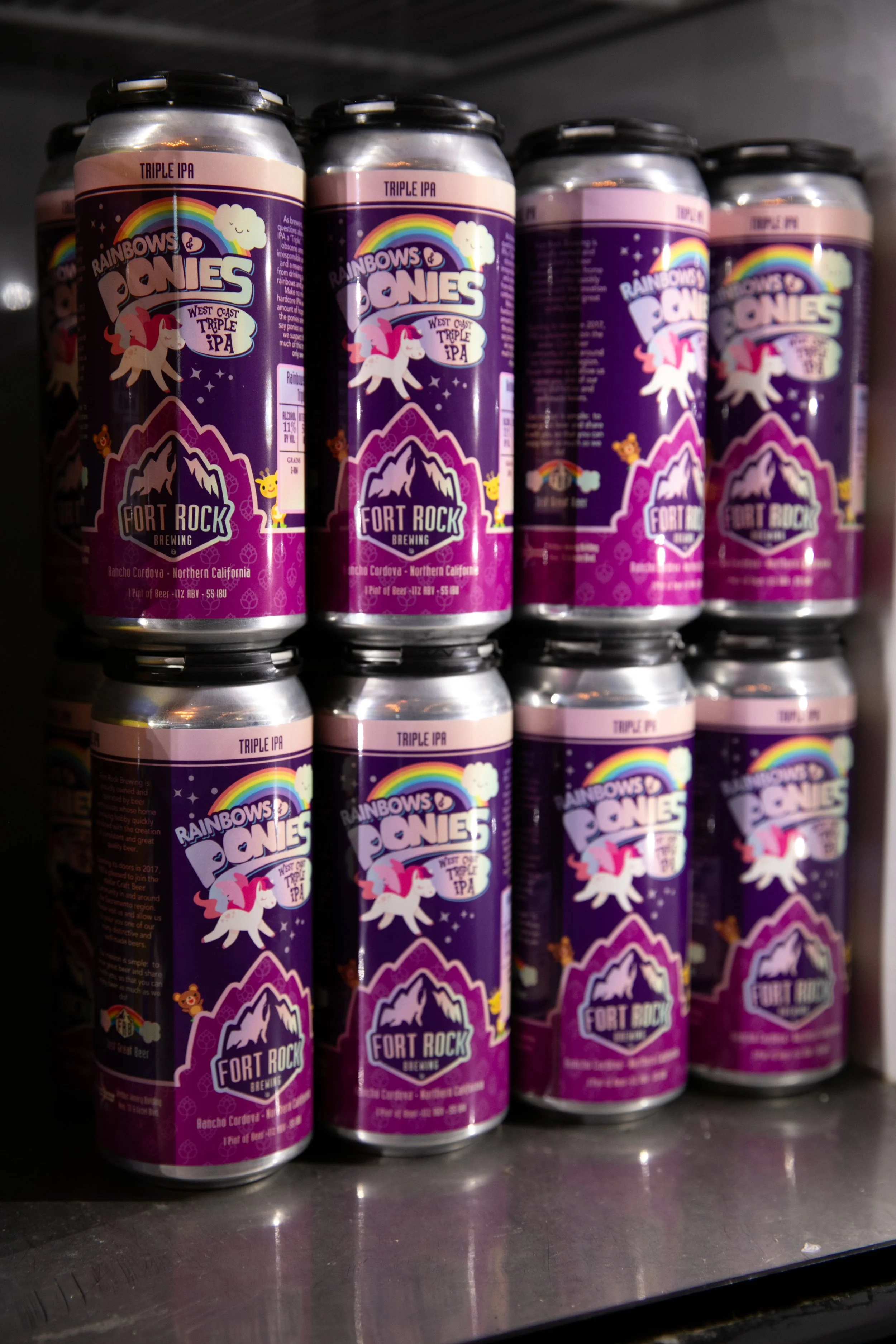 A display of cans of Rainbow Ponies West Coast Triple IPA beer by Fort Rock Brewing, featuring a purple label with a rainbow, clouds, and a cartoon pony.