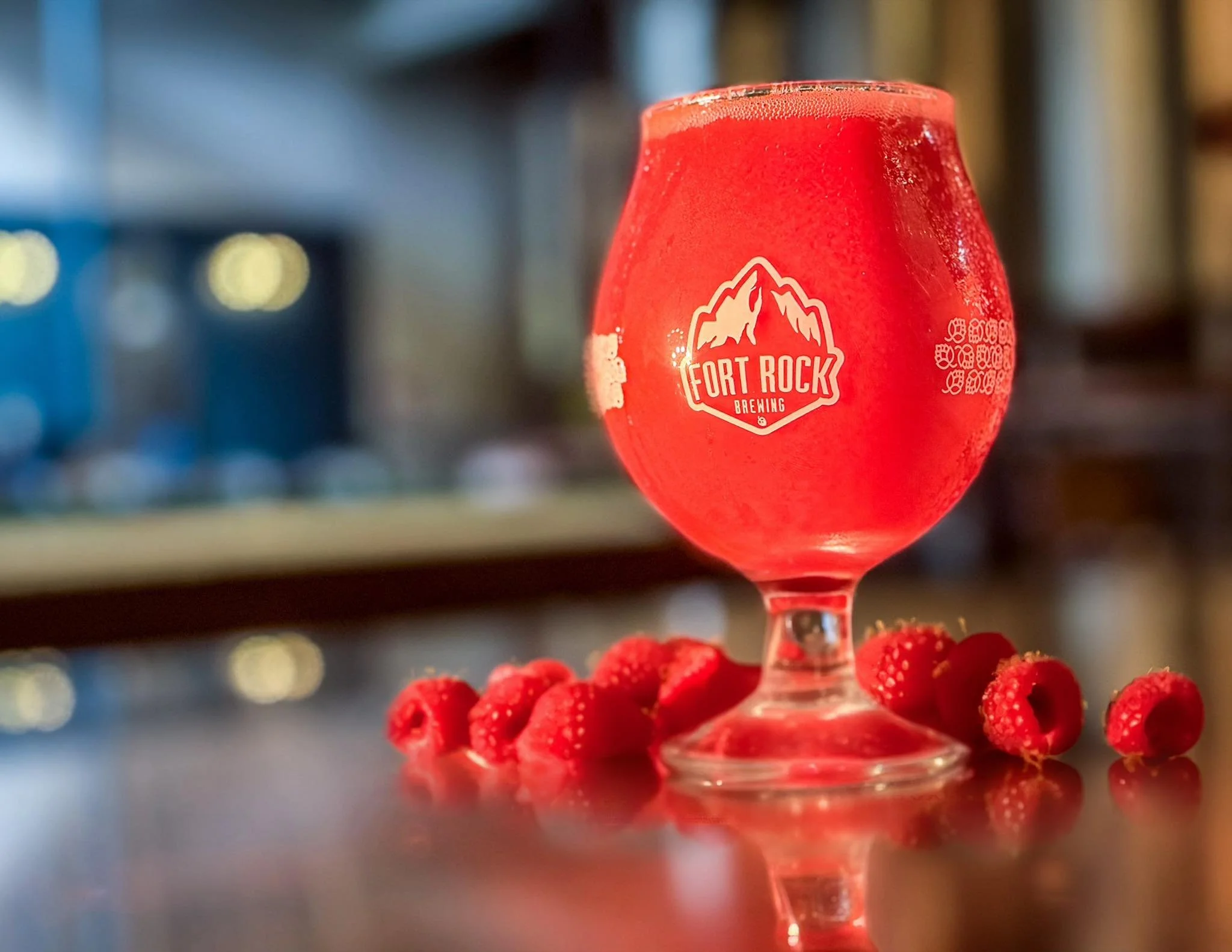 A red drink in a tulip-shaped glass with a "Fort Rock Brewing" logo, surrounded by red raspberries, on a reflective surface.