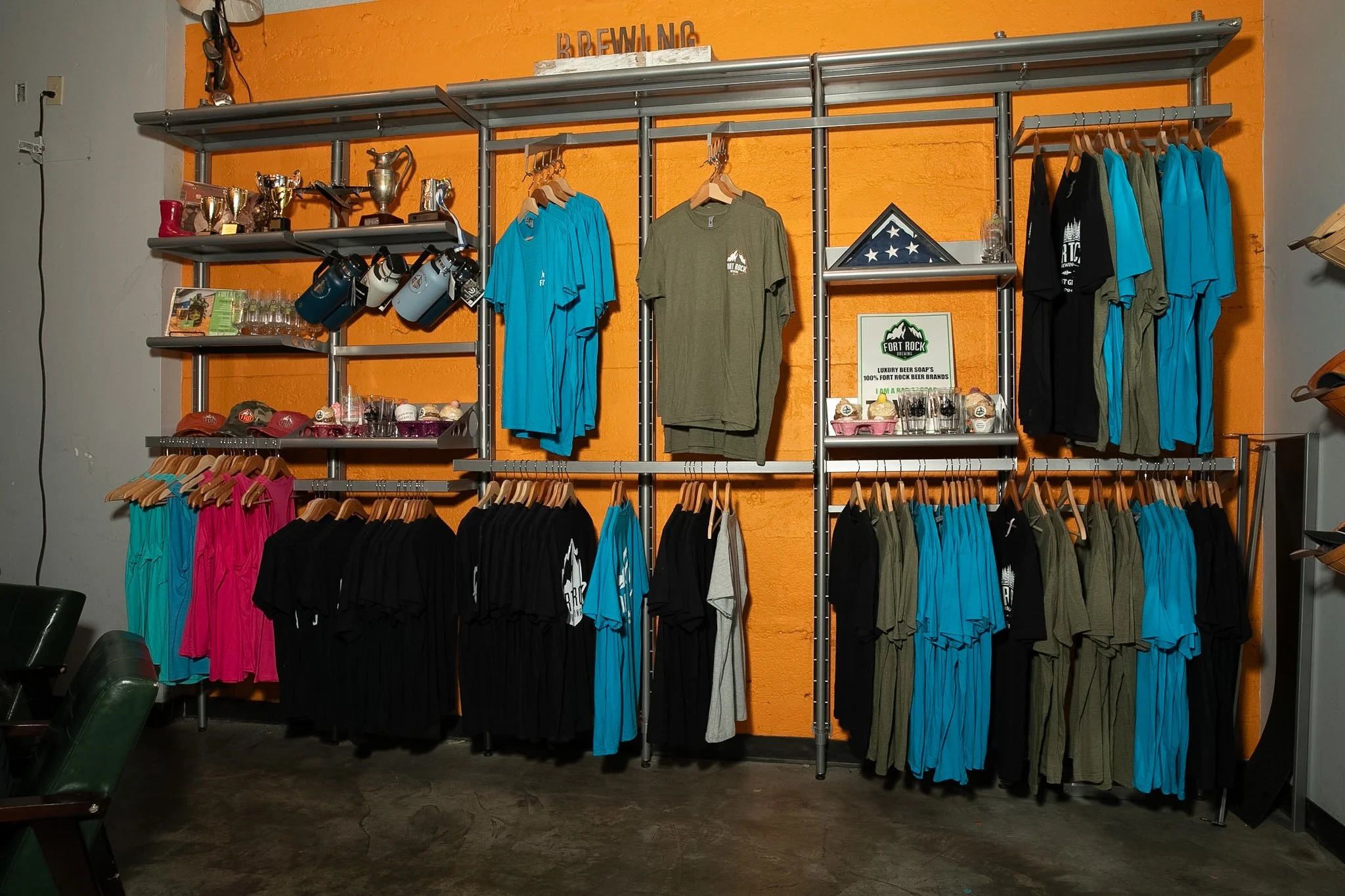 Clothing display rack with T-shirts, hats, trophies, and mugs in a room with orange and gray walls.