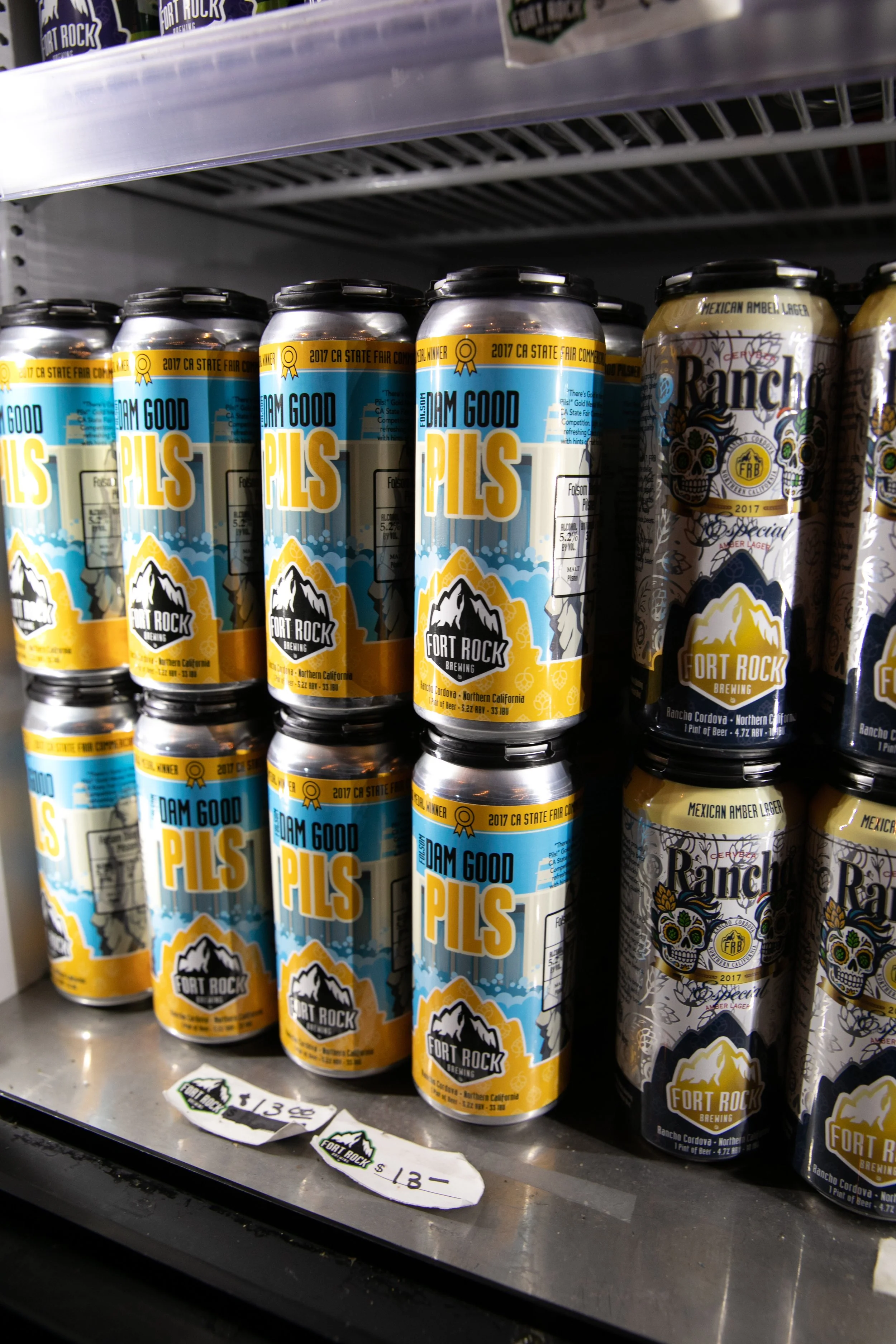 Rows of canned beer on a store shelf, including 'Dream Good Pils' from Fort Rock Brewing and 'Ranch' Mexican Amber Lager, with prices noted as $13 and $18.