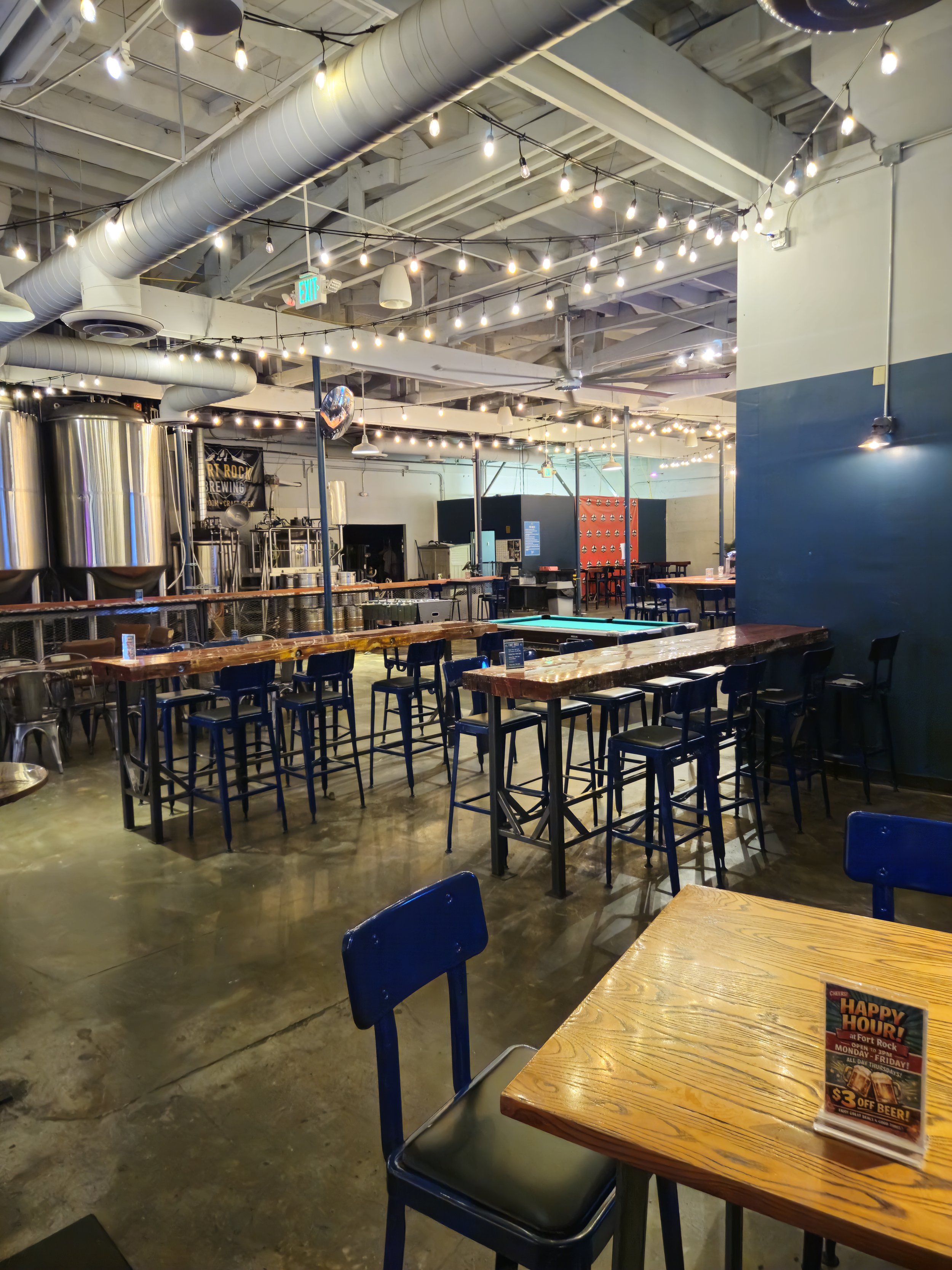 Interior of a brewery or bar with wooden tables and blue chairs, string lights hanging from the ceiling, large silver brewing tanks in the background, and a small promotional sign on a table advertising happy hour.