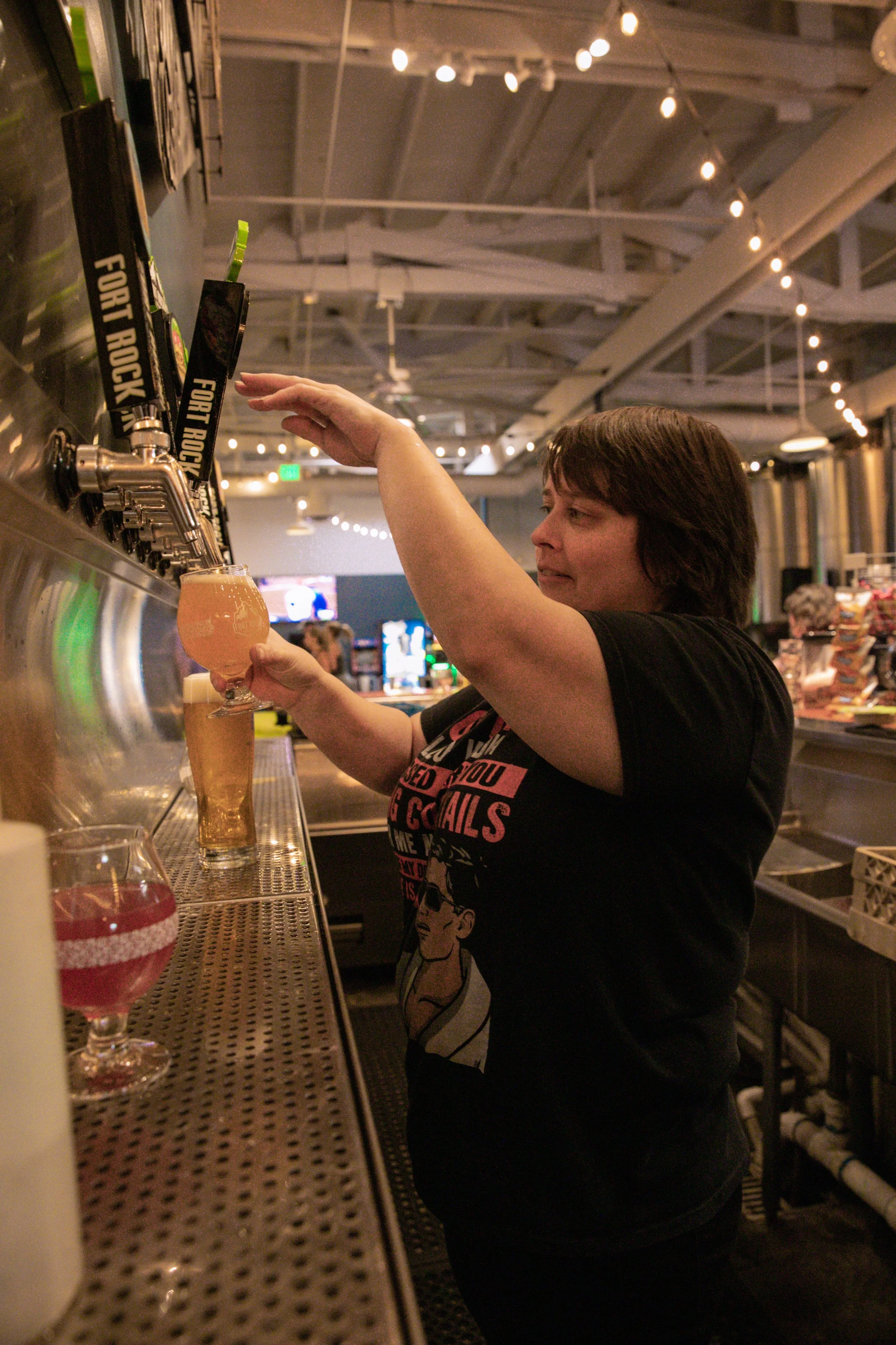 A woman with short dark hair pouring a glass of beer at a bar. She is wearing a black T-shirt and is standing in front of beer taps labeled 'FOR RACK' in a dimly lit bar or brewery.