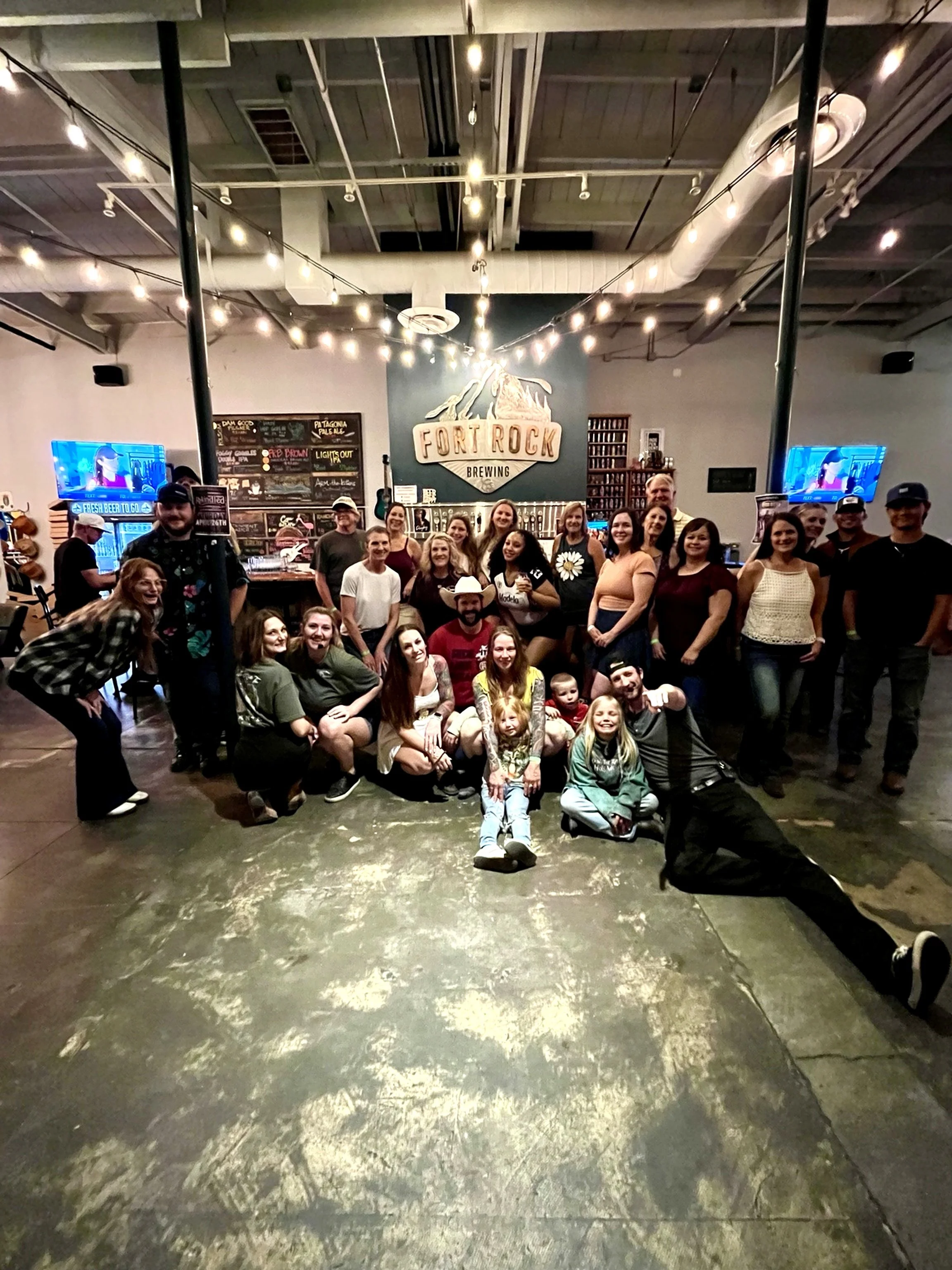 A large group of people posing and smiling for a photo inside a brewery, with the brewery's sign 'Fort Rock Brewing' in the background.