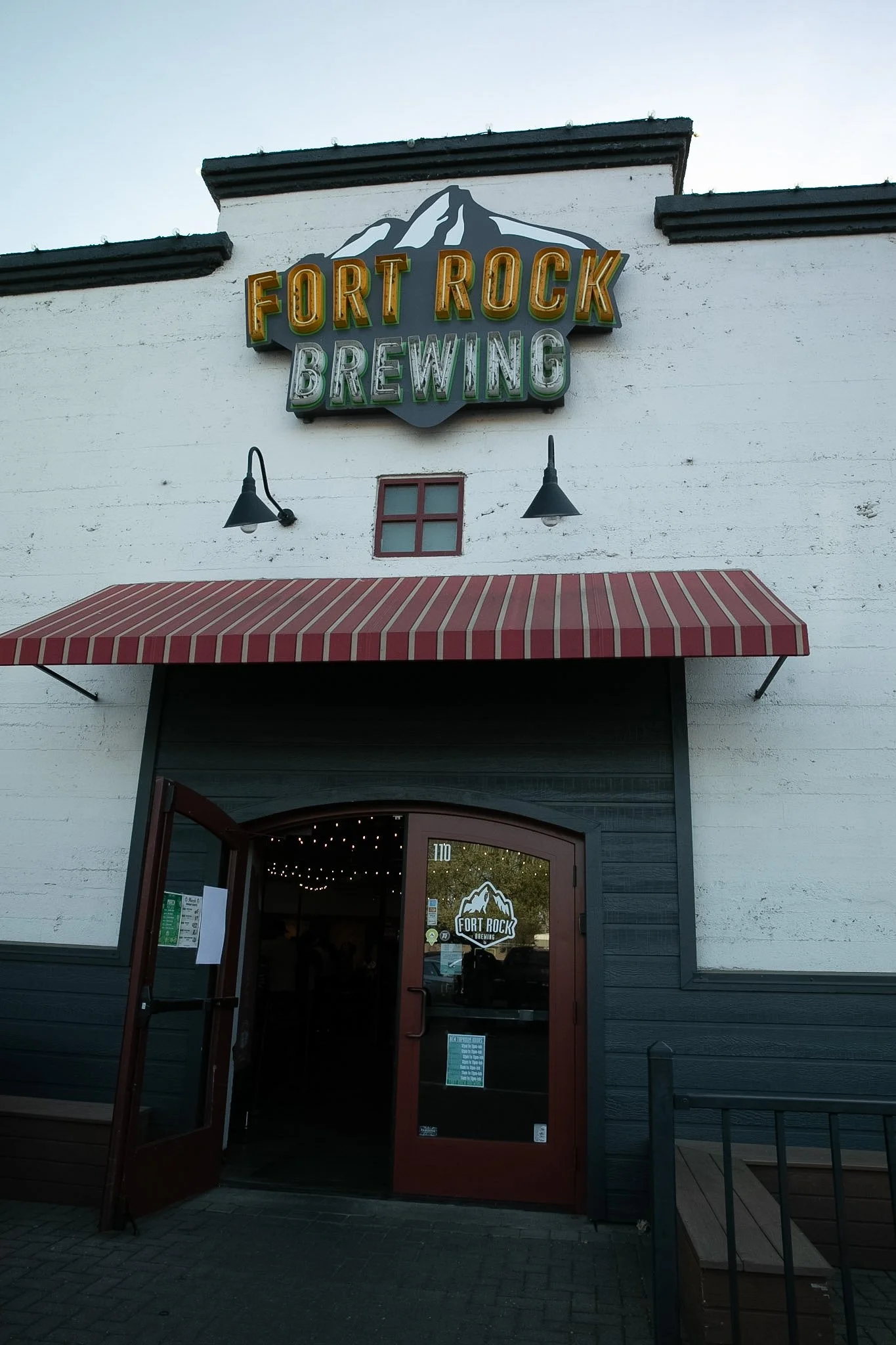 Exterior of Fort Rock Brewing restaurant with a sign featuring a mountain and snow, red and white striped awning, and dark-colored door.