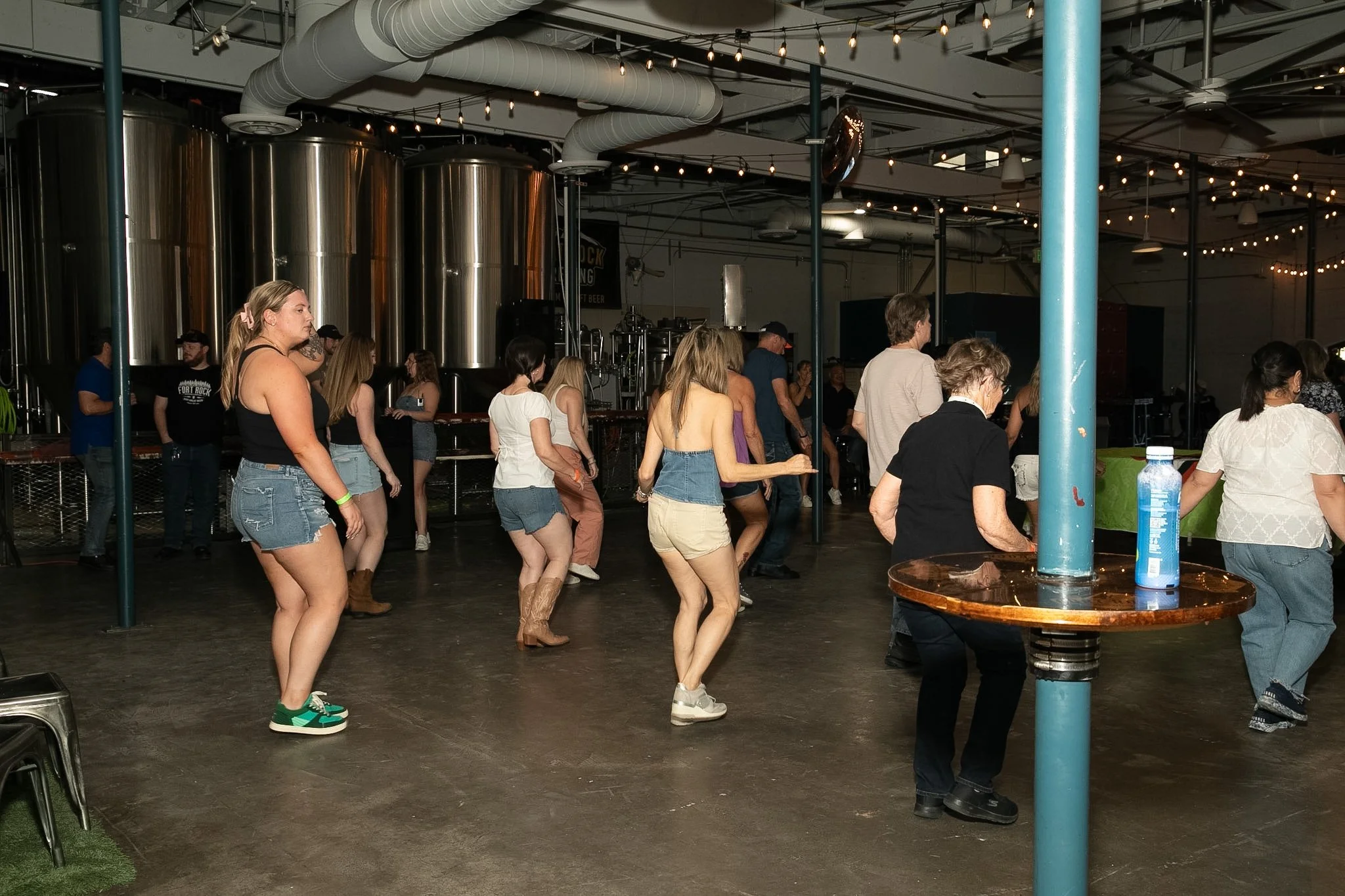 People line dancing inside a brewery or event space with large industrial brewing tanks in the background. There are string lights overhead.