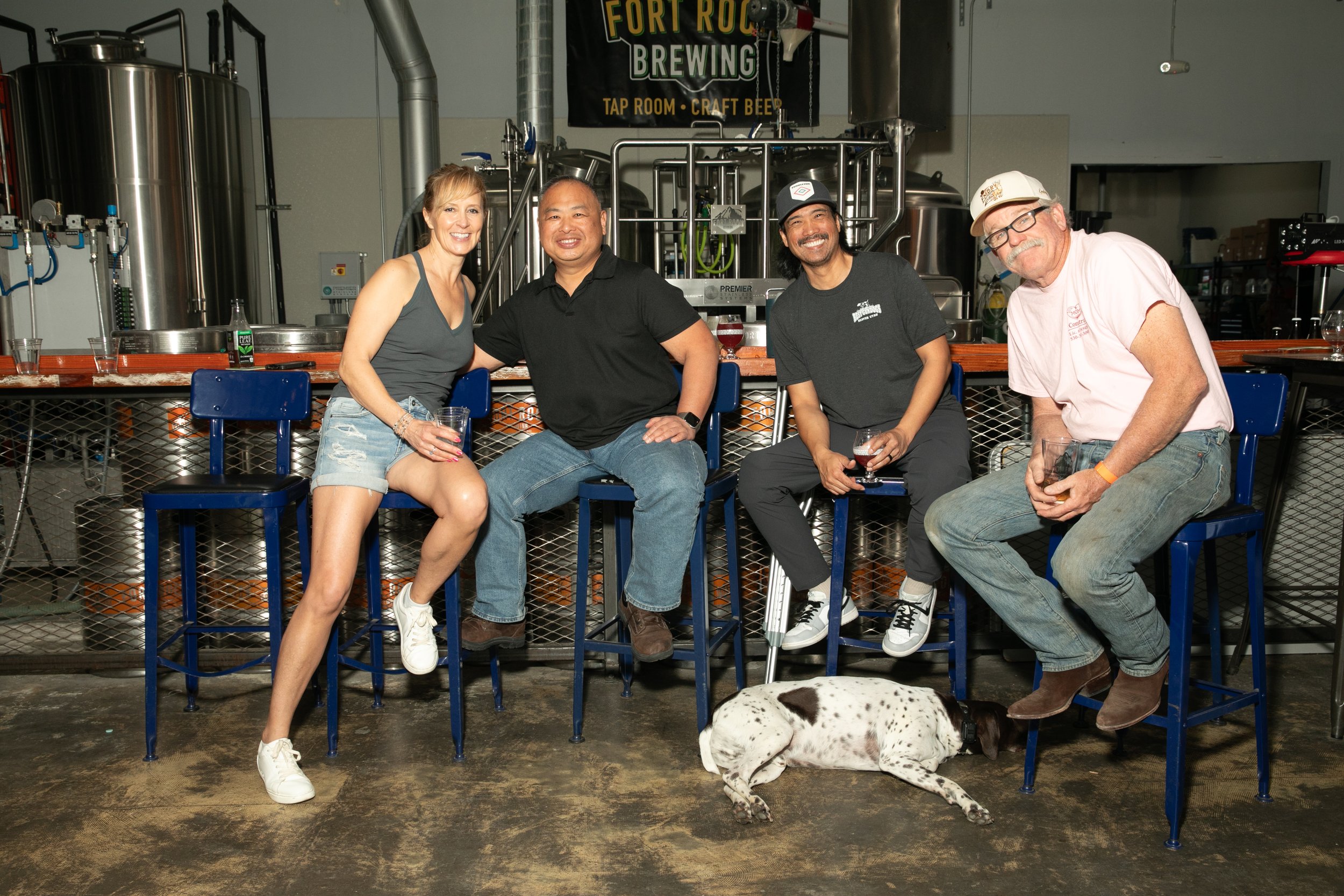 Four people enjoying drinks at a brewery bar, with brewing equipment in the background and a Dalmatian dog lying on the floor in front.