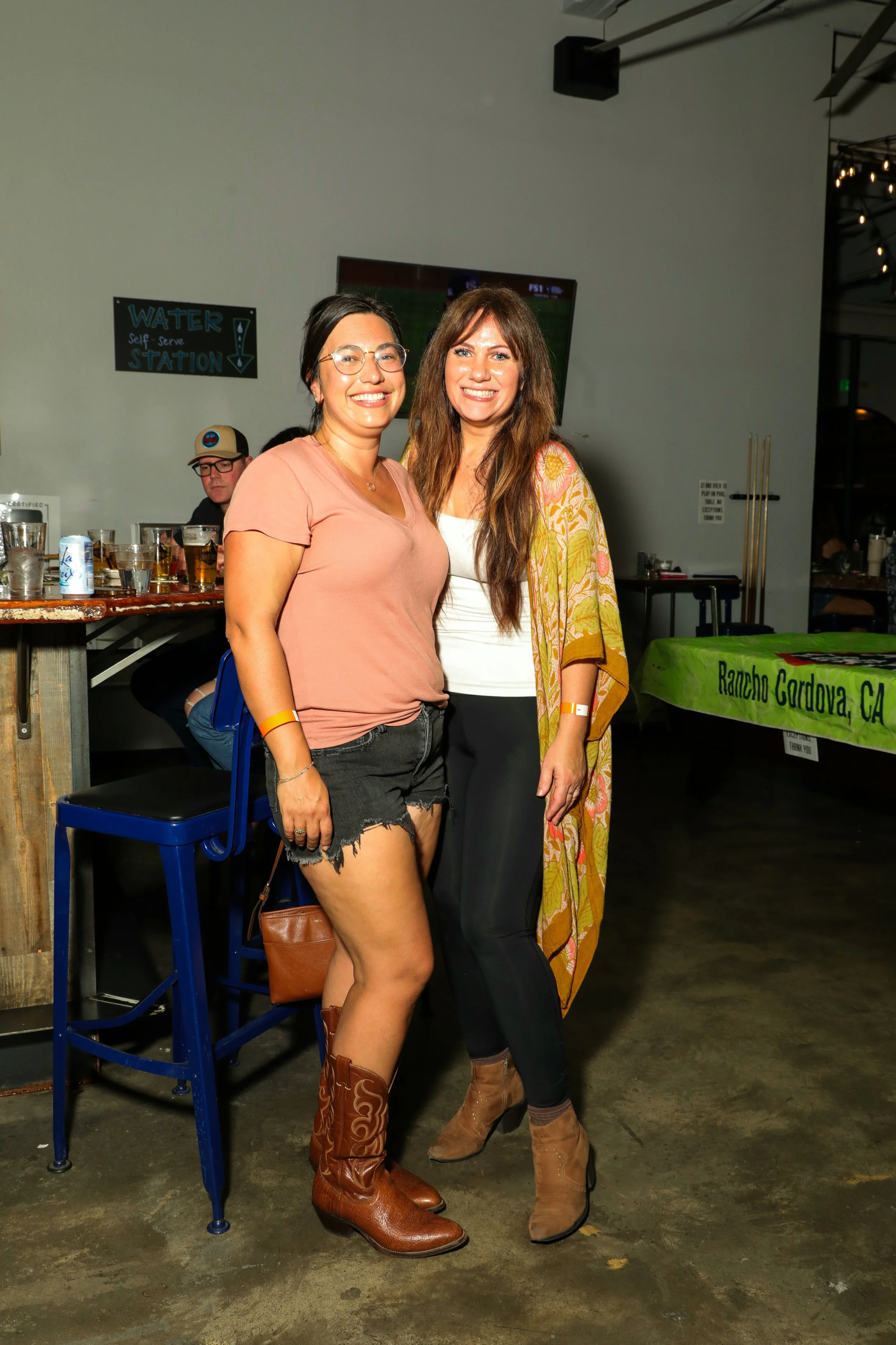 Two women smiling and posing together at an indoor event. One is wearing a peach-colored t-shirt, black denim shorts, and cowboy boots, while the other is dressed in a white top, black pants, and tan boots. There is a man behind them at a bar, and a 