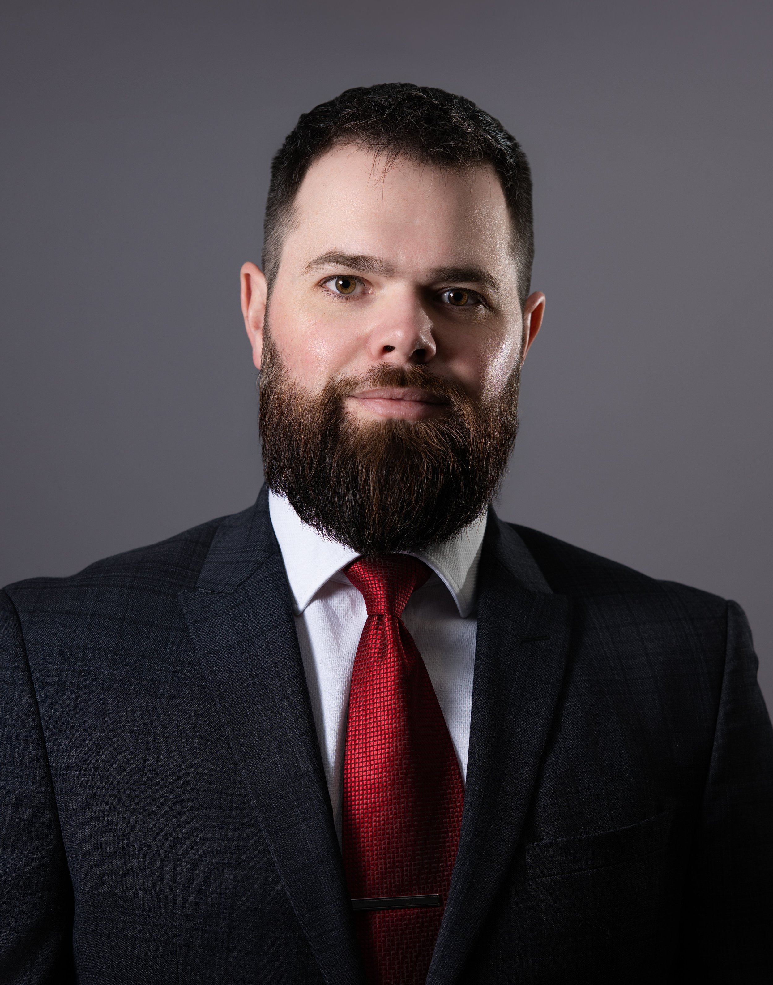 Professional man in a suit with a red tie and beard, posing against a gray background.