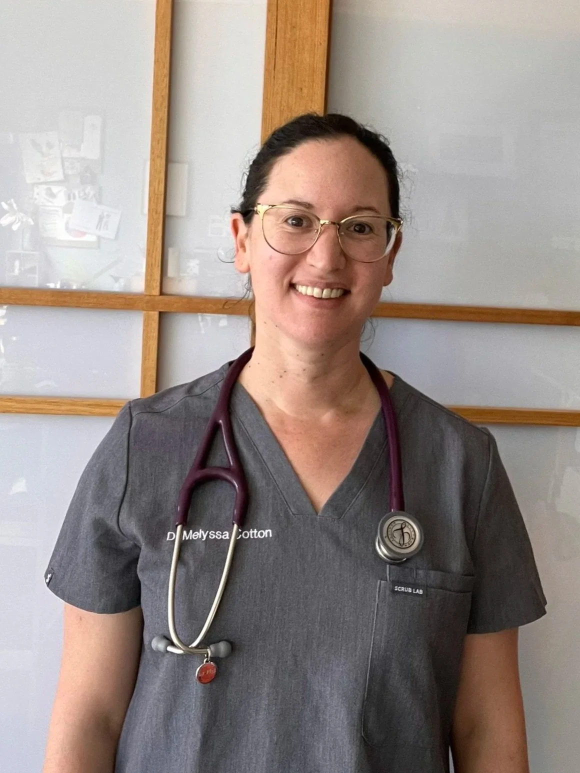 A smiling woman wearing glasses and medical scrubs with a stethoscope around her neck, standing indoors in front of a wall with a wooden grid.