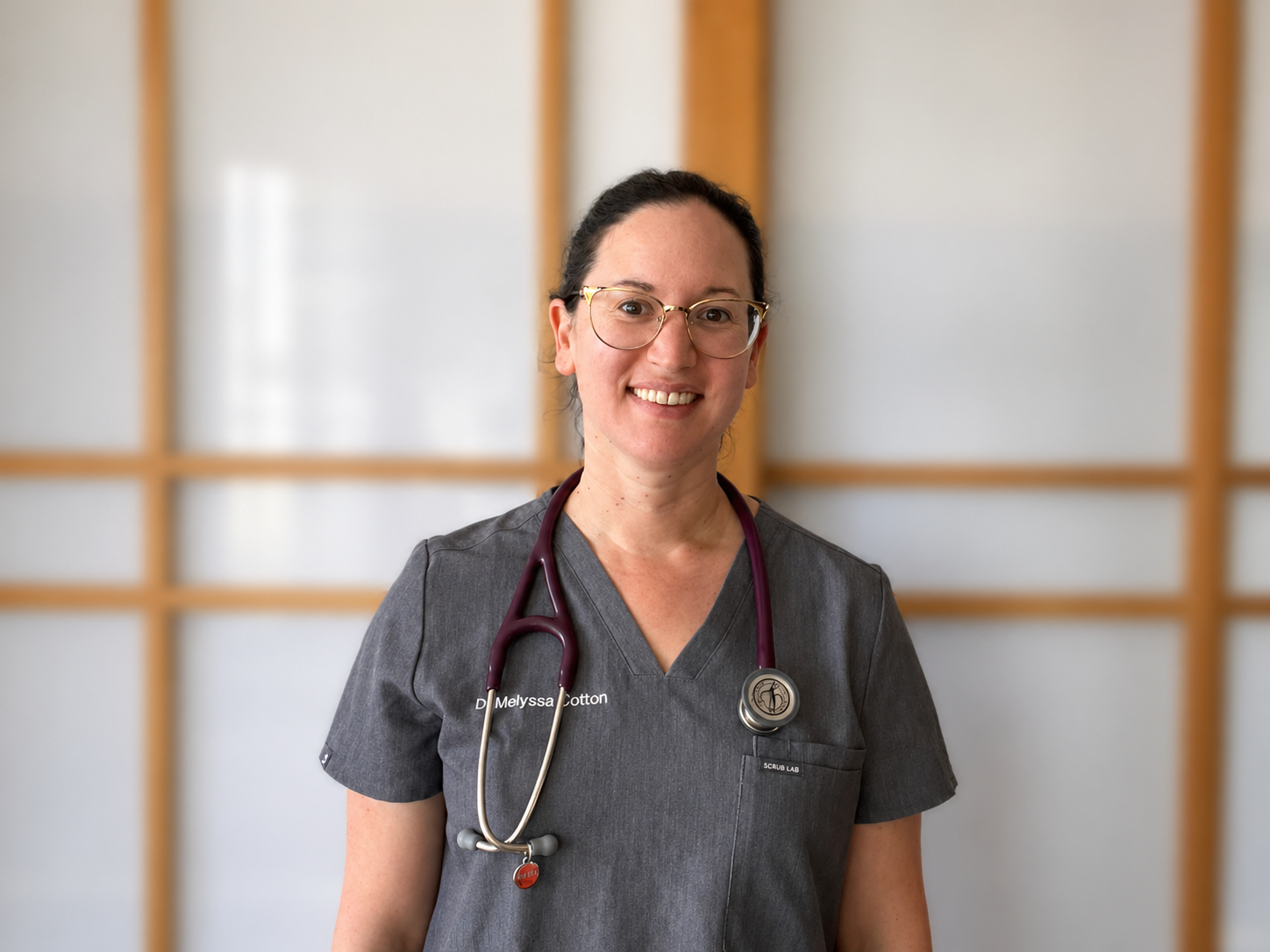 A smiling female mobile vet wearing glasses and medical scrubs with a stethoscope around her neck, smiling at the camera in a warm and friendly way.