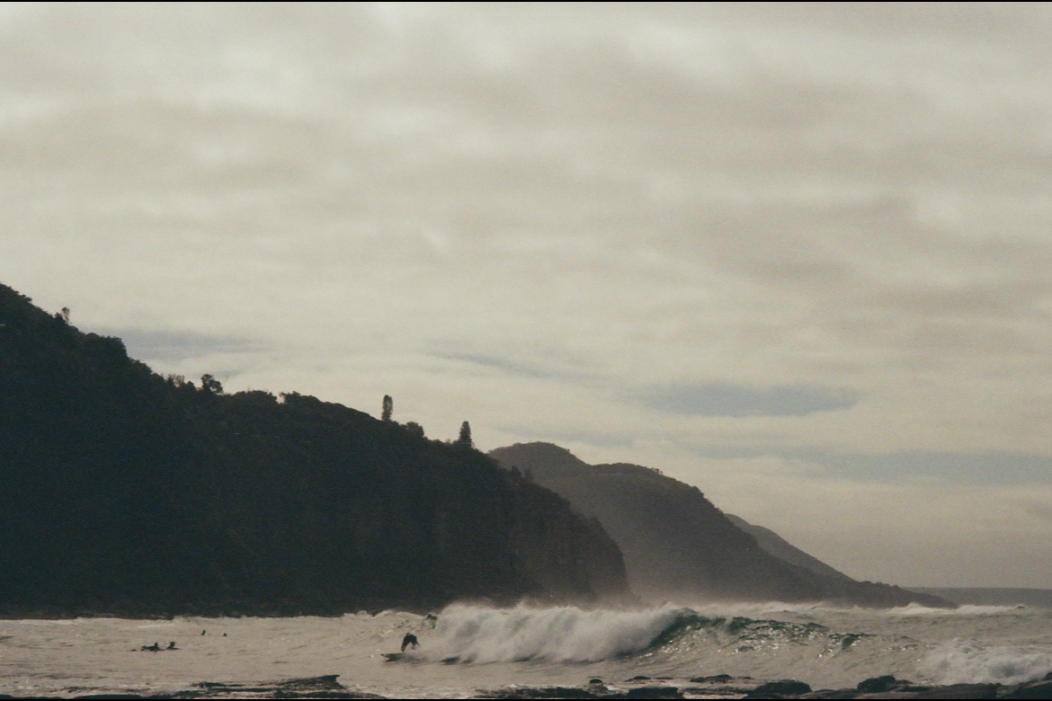 Beach scene with surfers in the water, ocean waves, and a distant hilly coastline under an overcast sky.