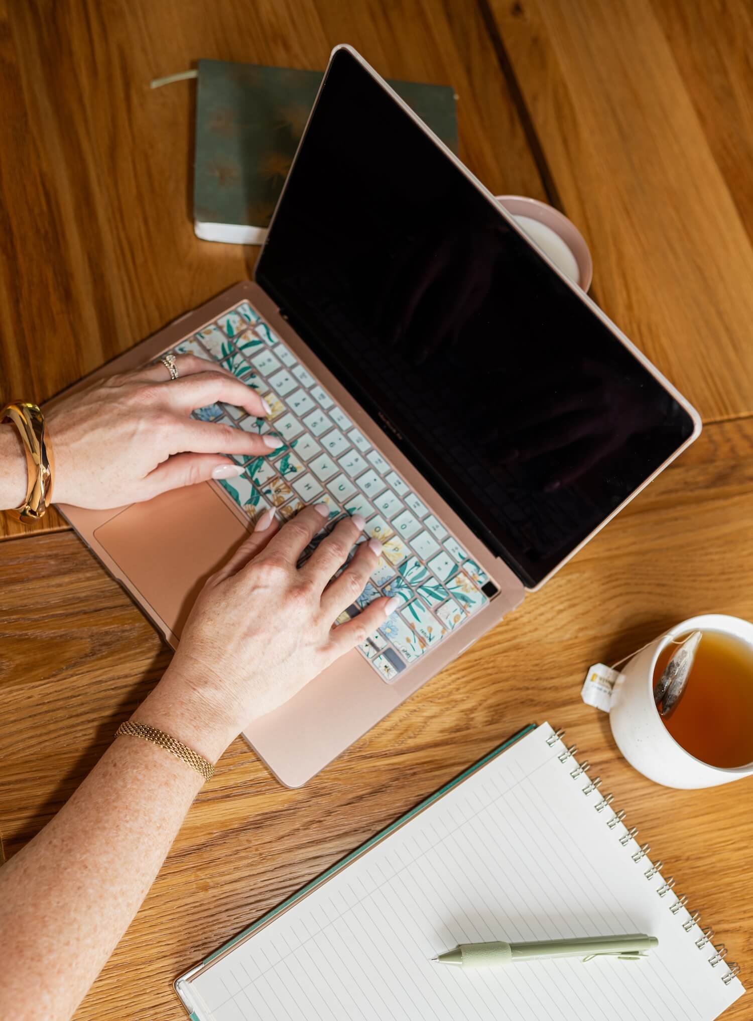 Hands typing on a laptop at a wooden table with a notebook, pen, and a cup of tea nearby.