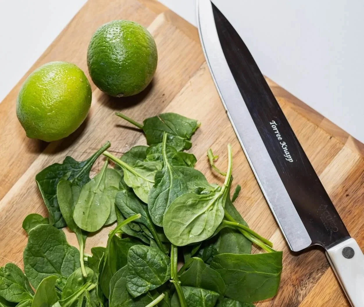 Fresh spinach leaves and two whole limes on a wooden cutting board next to a kitchen knife.