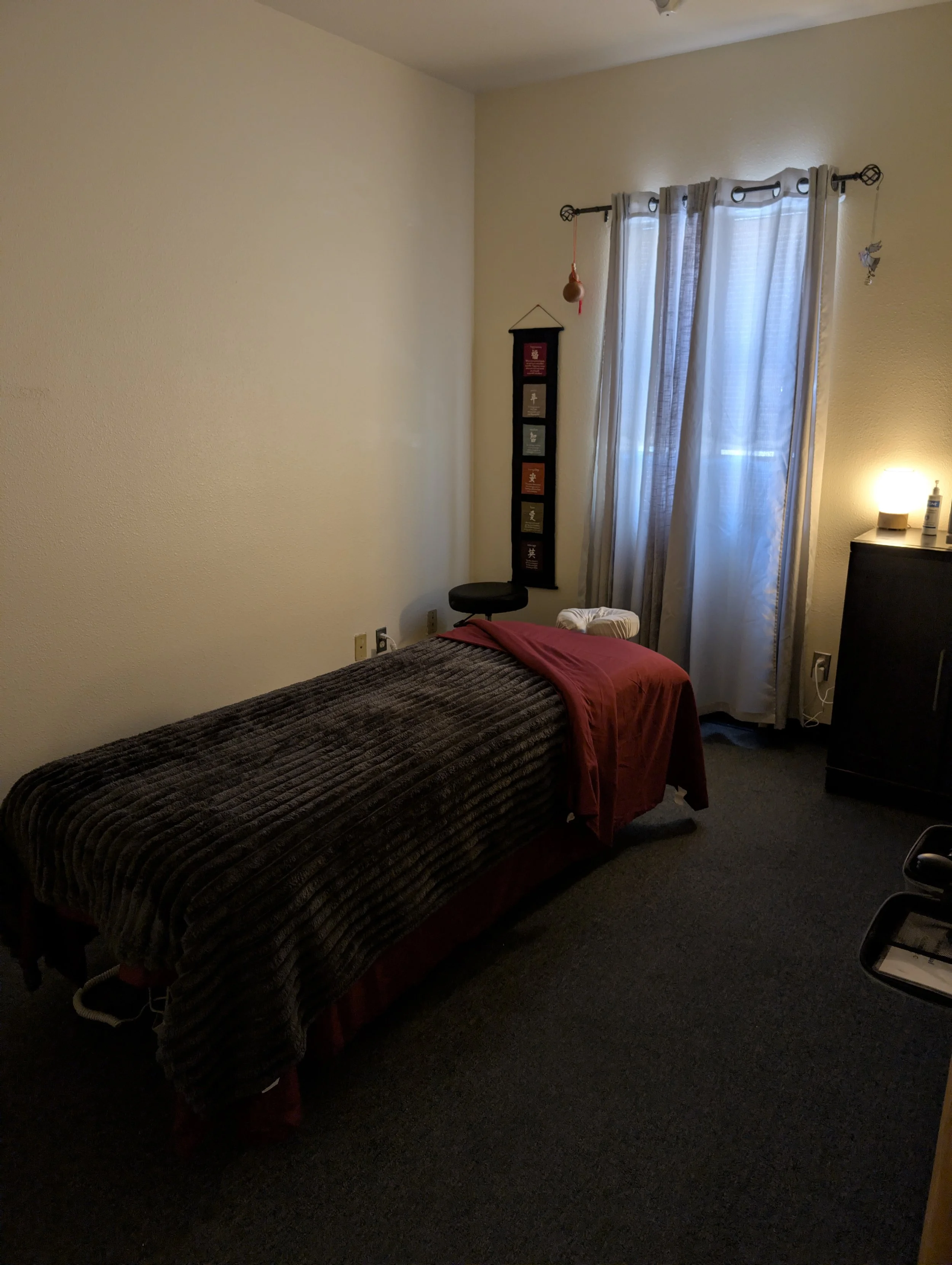 A bedroom with a bed covered with a dark brown blanket and a red sheet, a small black stool against the wall, a wall hanging with colorful sections, a window with white curtains, a small dresser with a lamp and bottle of lotion, and a black office chair.