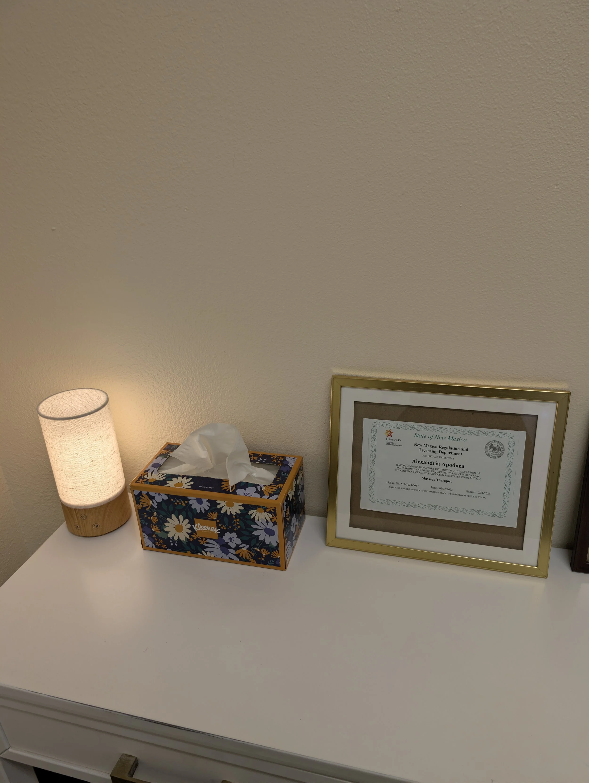 A table with a lit lamp, a box of tissues, and a framed certificate on the wall.