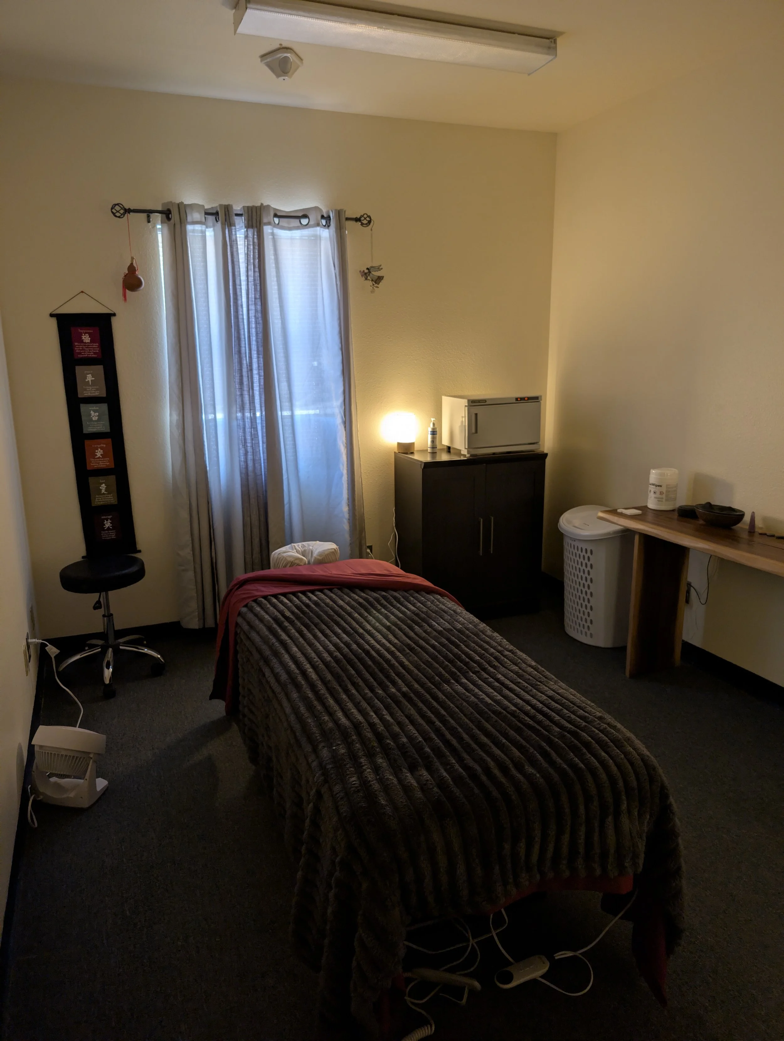 A bedroom with beige walls, a bed covered in a brown and gray striped cover, and a window with white curtains. There is a small black table with an orange hanging ornament, a lamp, a microwave on a dark cabinet, a table with some items including wipes, and a laundry basket. A small hanging decoration and a vertical wall hanging with motivational quotes are visible.