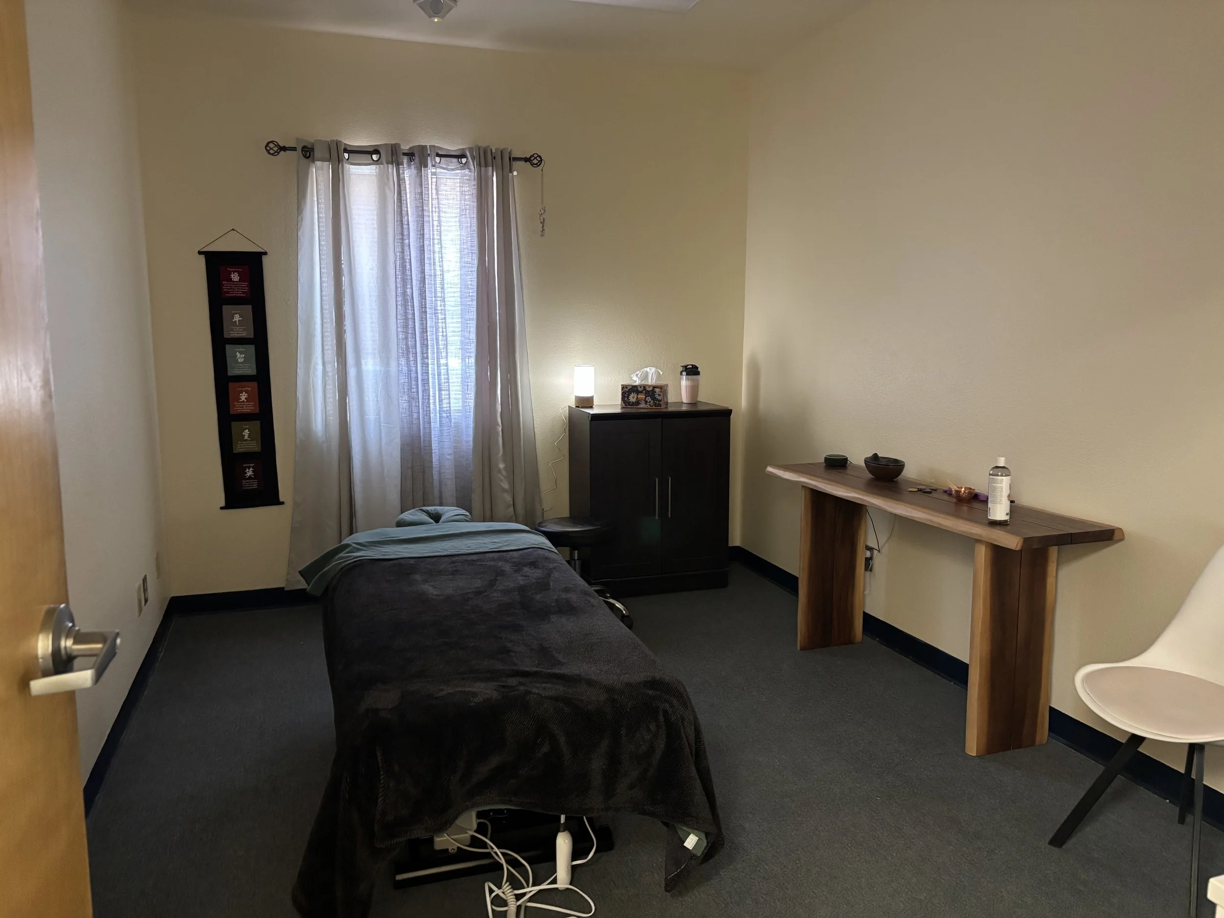 A bedroom with a bed, a window with sheer curtains, a tall black cabinet, a small table with a tissue box, water bottle, and a pink tumbler, and a small wooden desk with a black bowl, a bowl, and some electronic accessories.