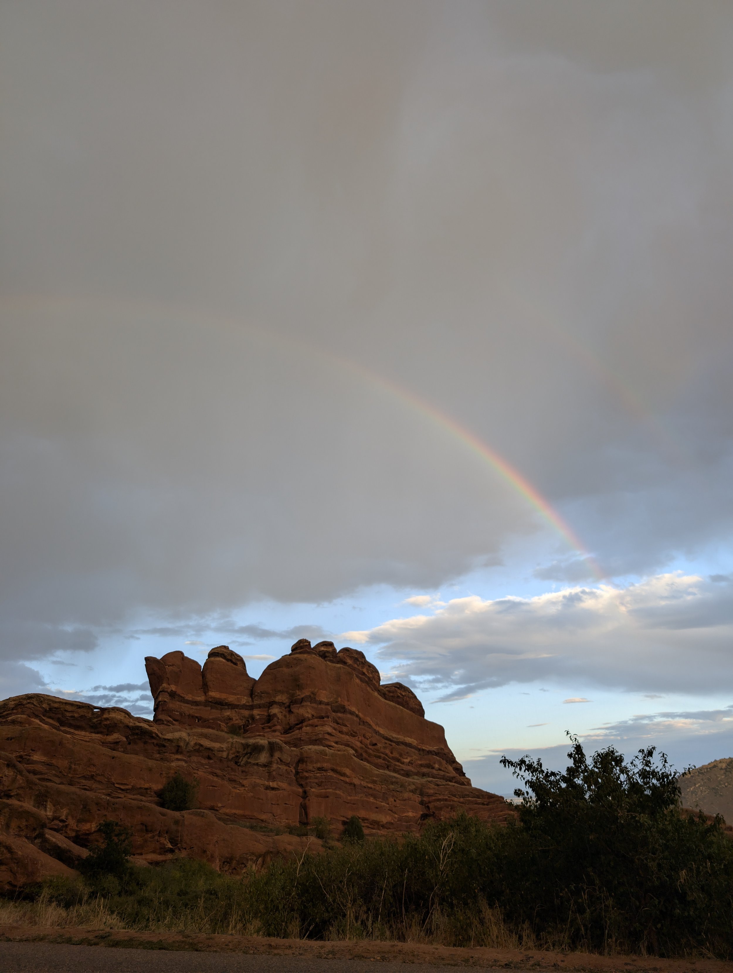 A landscape scene featuring reddish rocks and sparse vegetation in the foreground, with a double rainbow visible in the sky above, which is partly cloudy with blue sky patches.