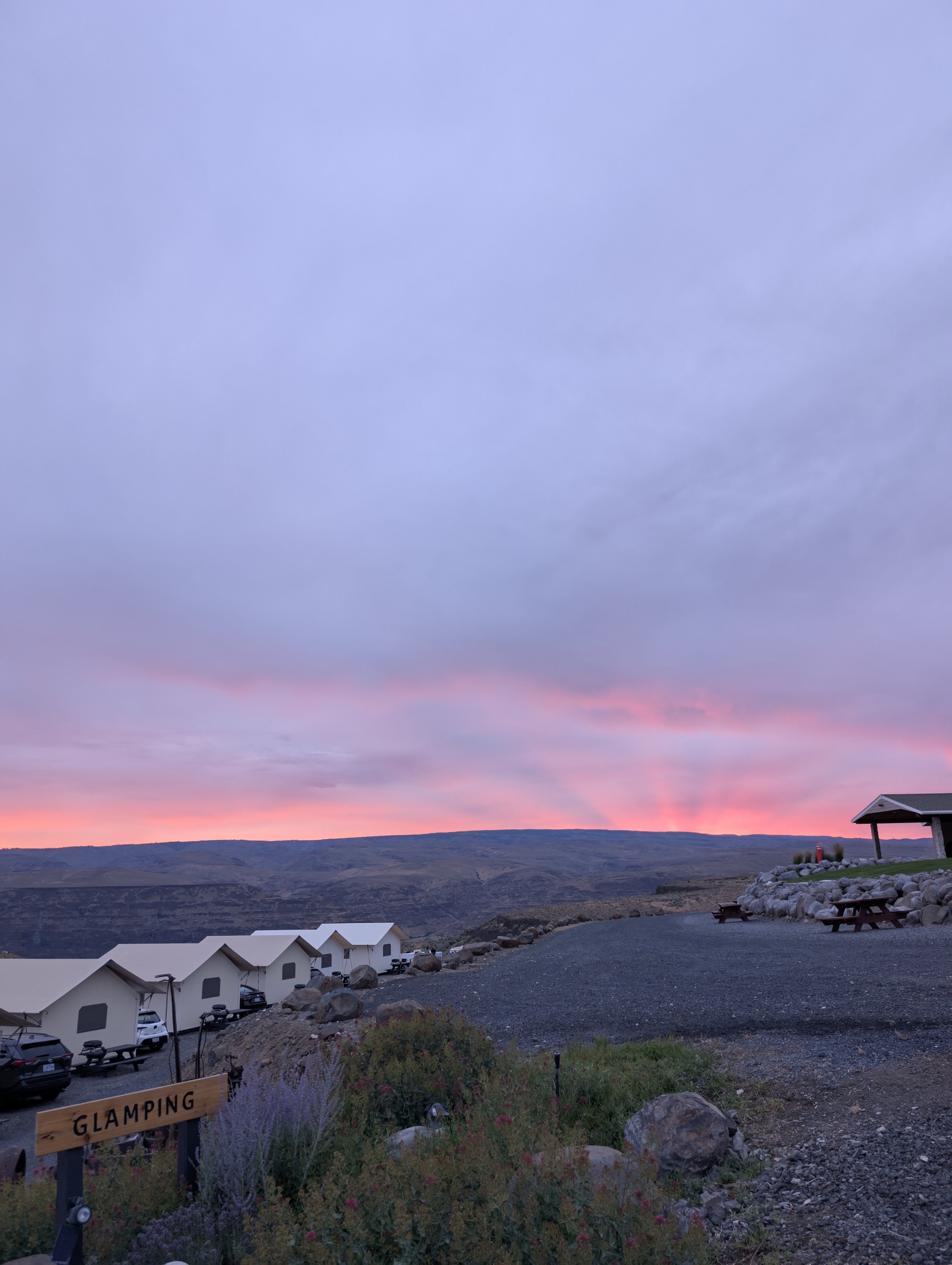 Landscape view of a sunset with pink and purple hues in the sky over a hilly area. There are tents with white roofs and parked cars at a glamping site.  There are some rocks, flowers, and a small pavilion on the right side.