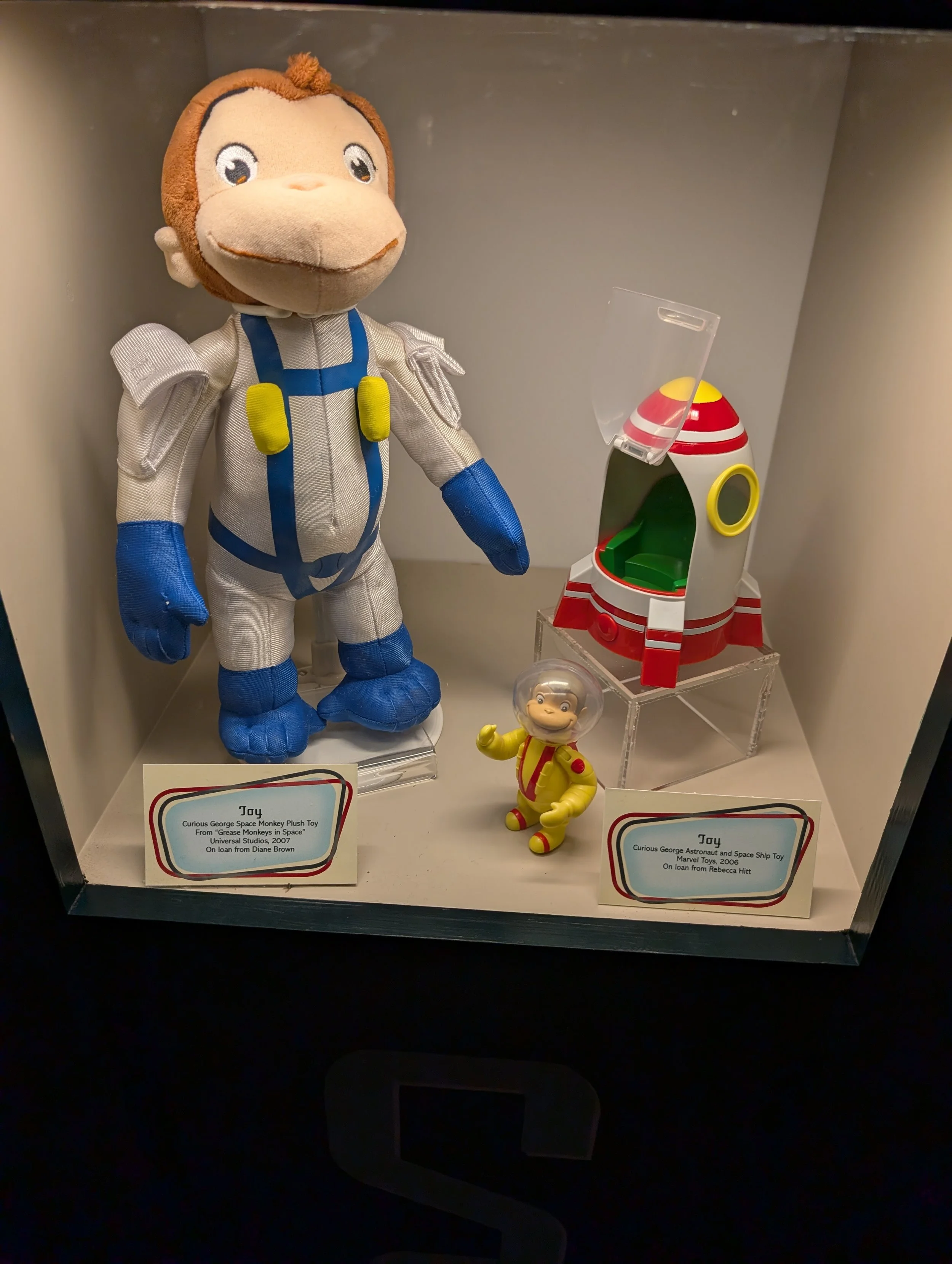 Two toys in a display case: a plush monkey dressed as an astronaut with a space suit and blue gloves, and a small plastic toy of a monkey in a yellow spacesuit. There are also two informational cards at the bottom describing the toys' names, origins, and year of creation.