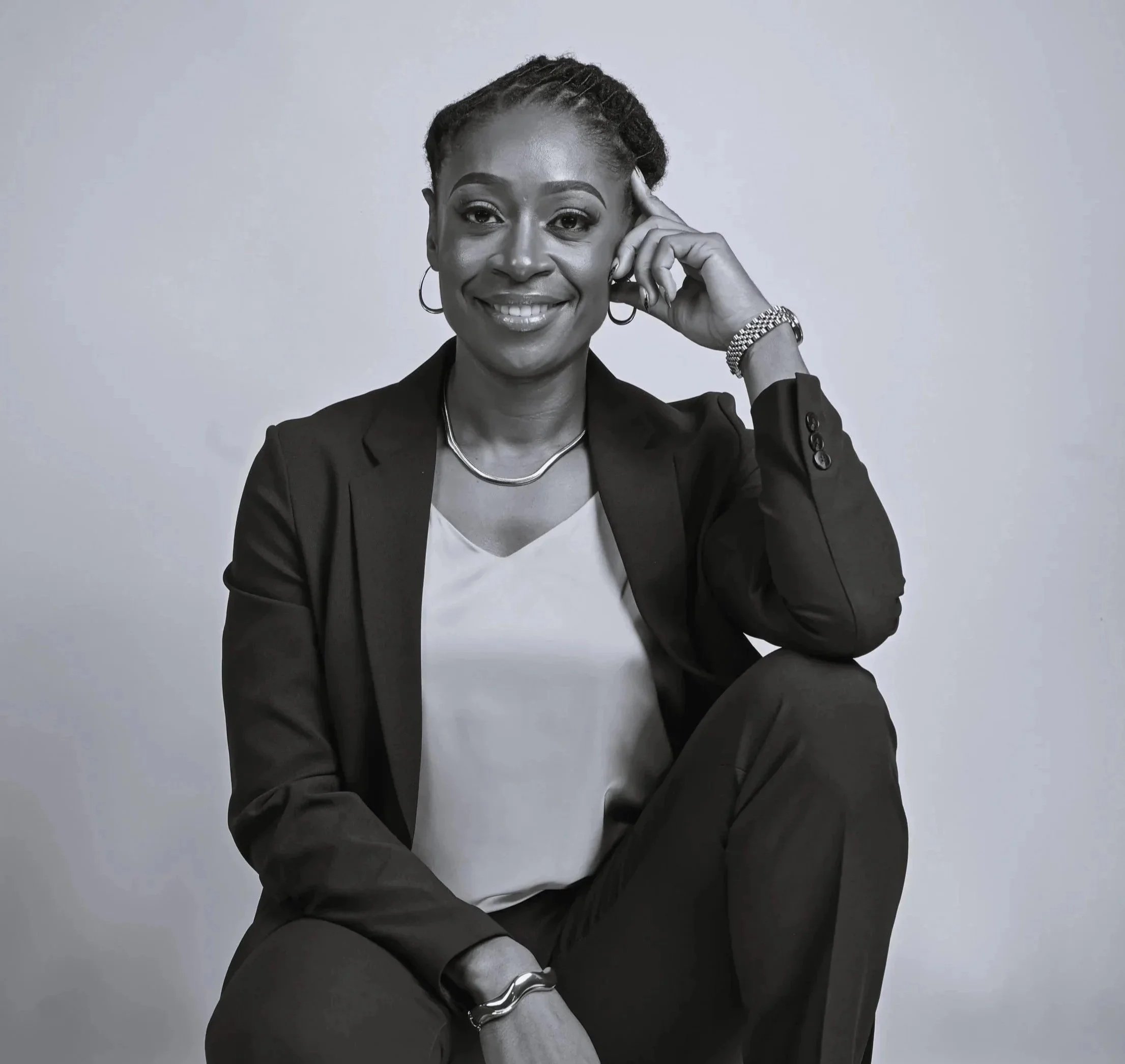 A woman in a black suit sitting against a plain background, smiling, with her right hand resting on her head.